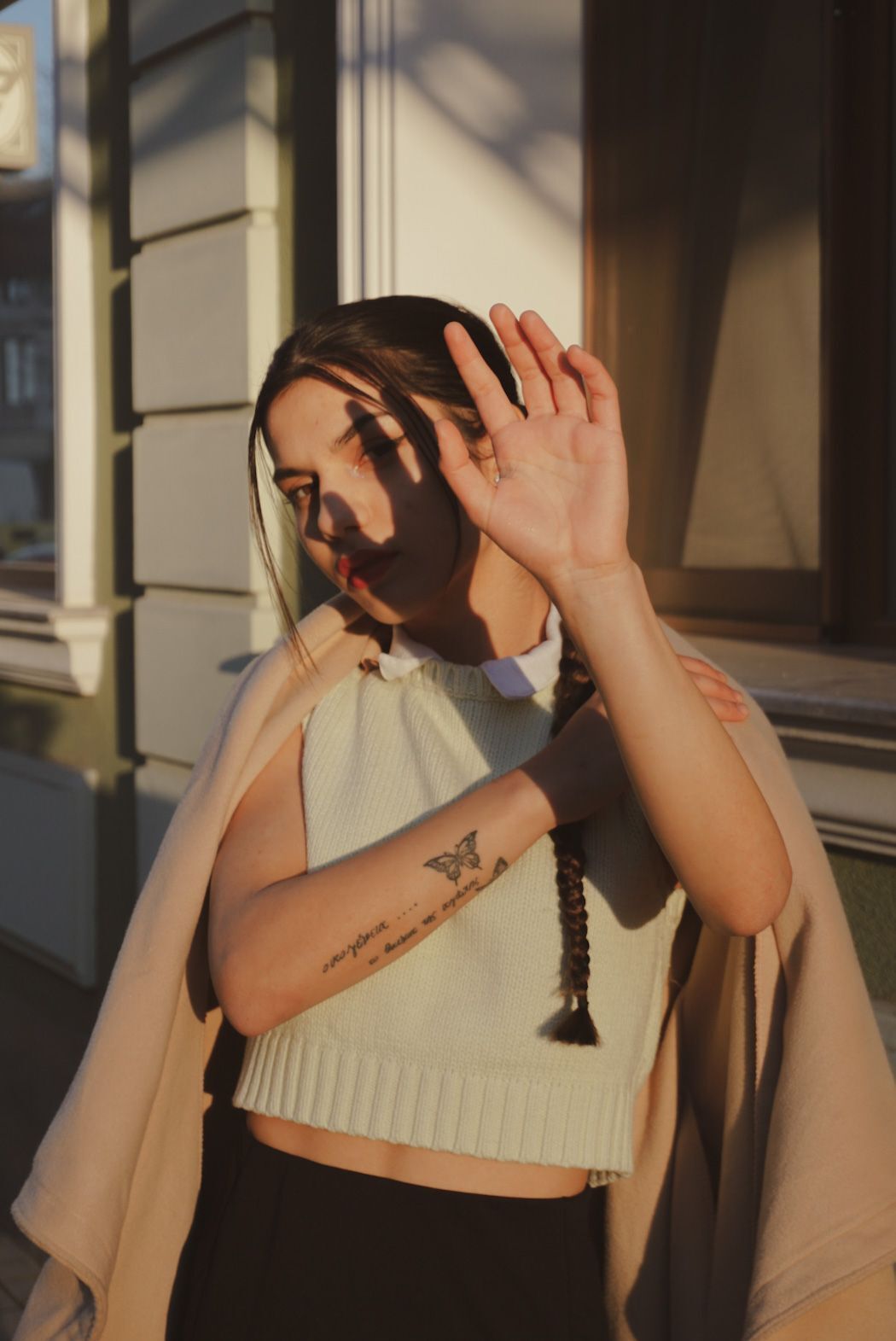 Young woman with braided hair shields her eyes from sunlight, showing butterfly tattoo on forearm, wearing white crop top outdoors.