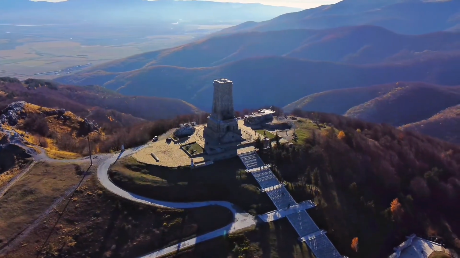 Aerial view of Shipka Monument atop mountain surrounded by autumn forest.