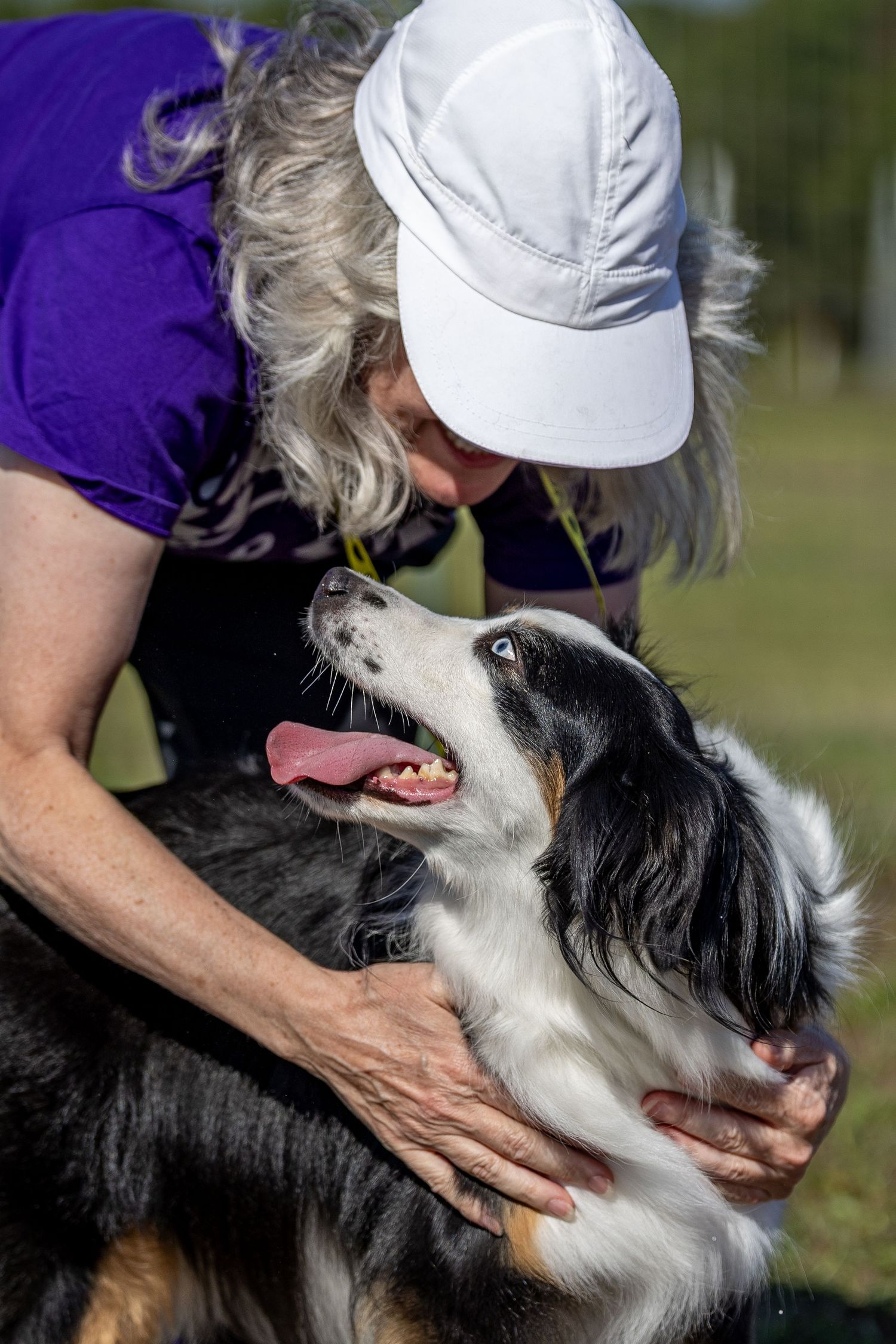 Woman in white cap and purple shirt lovingly embraces a tri-color Australian Shepherd dog outdoors.