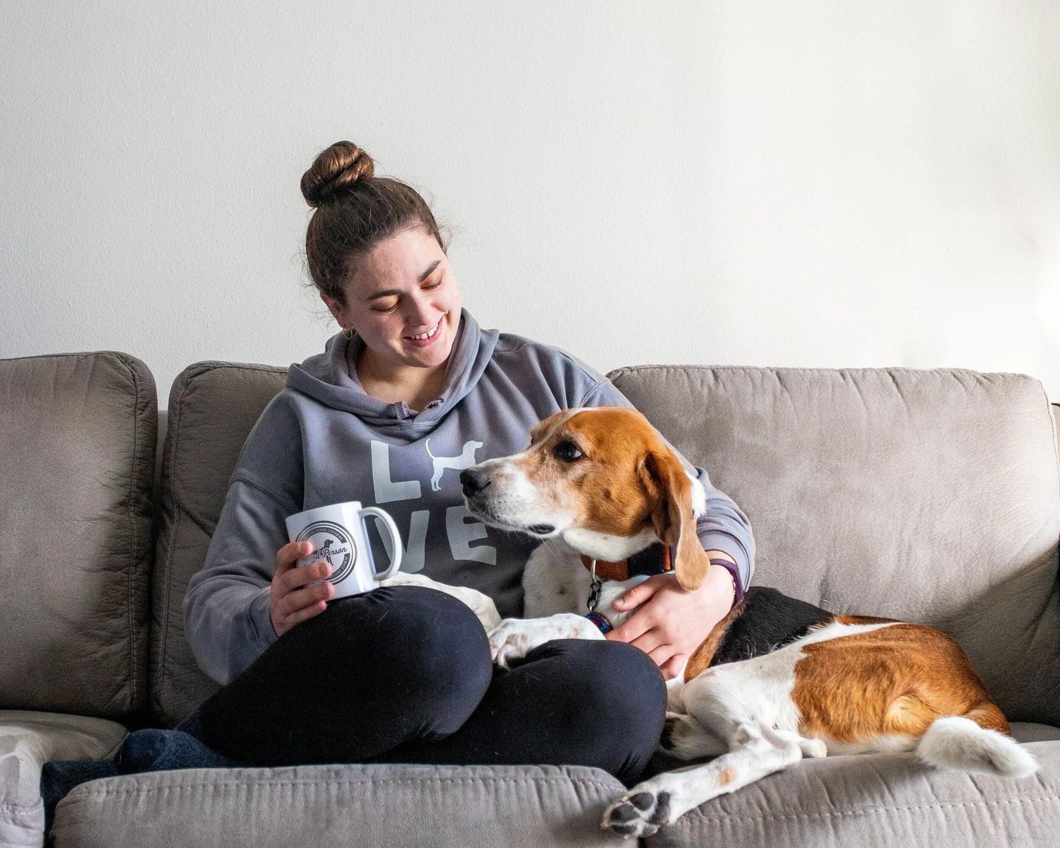 Young woman in gray hoodie smiling at beagle dog on gray couch while holding a white mug.