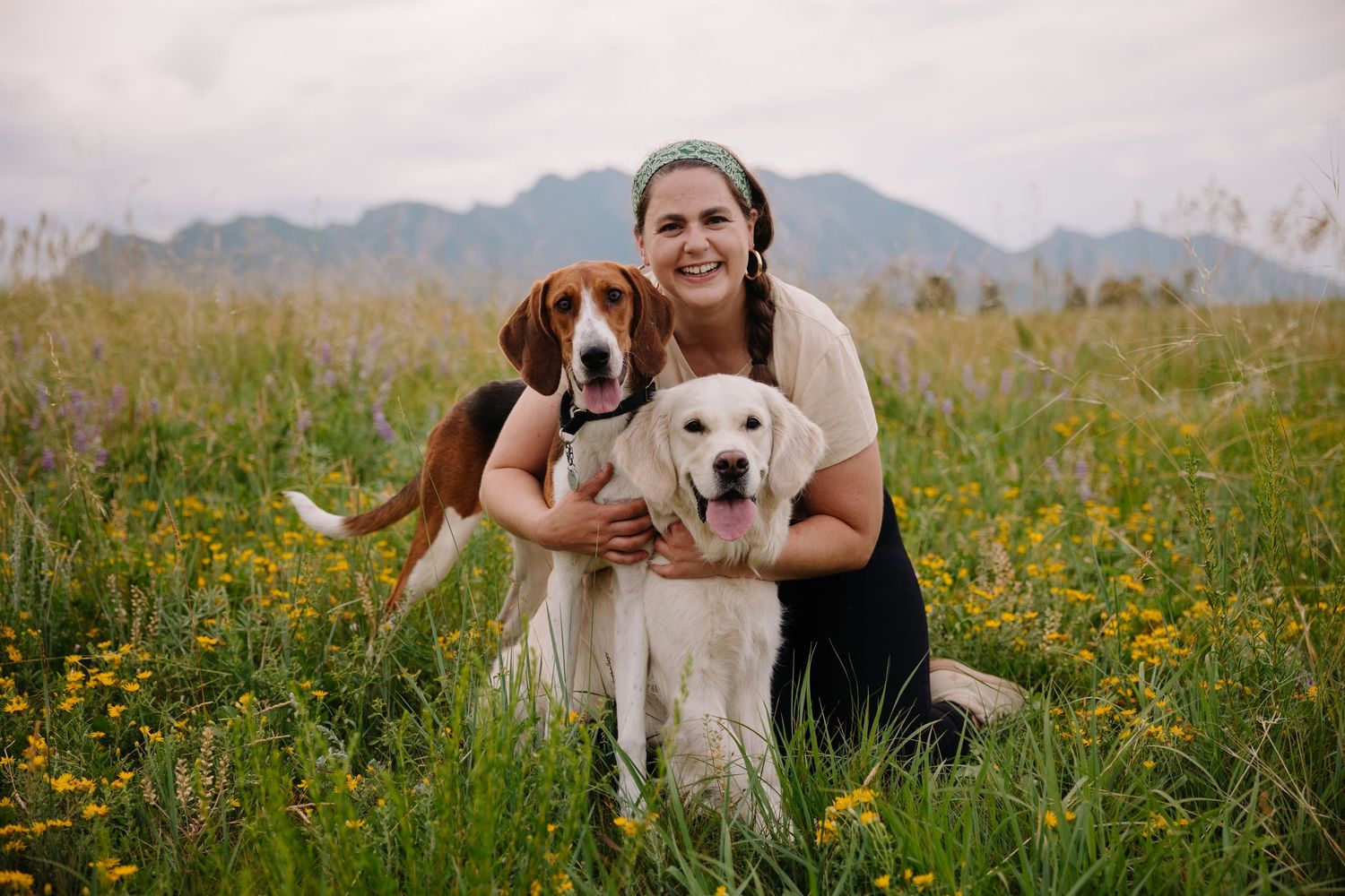 Woman smiling with two dogs, a beagle and yellow lab, in a wildflower meadow with mountains in the background.