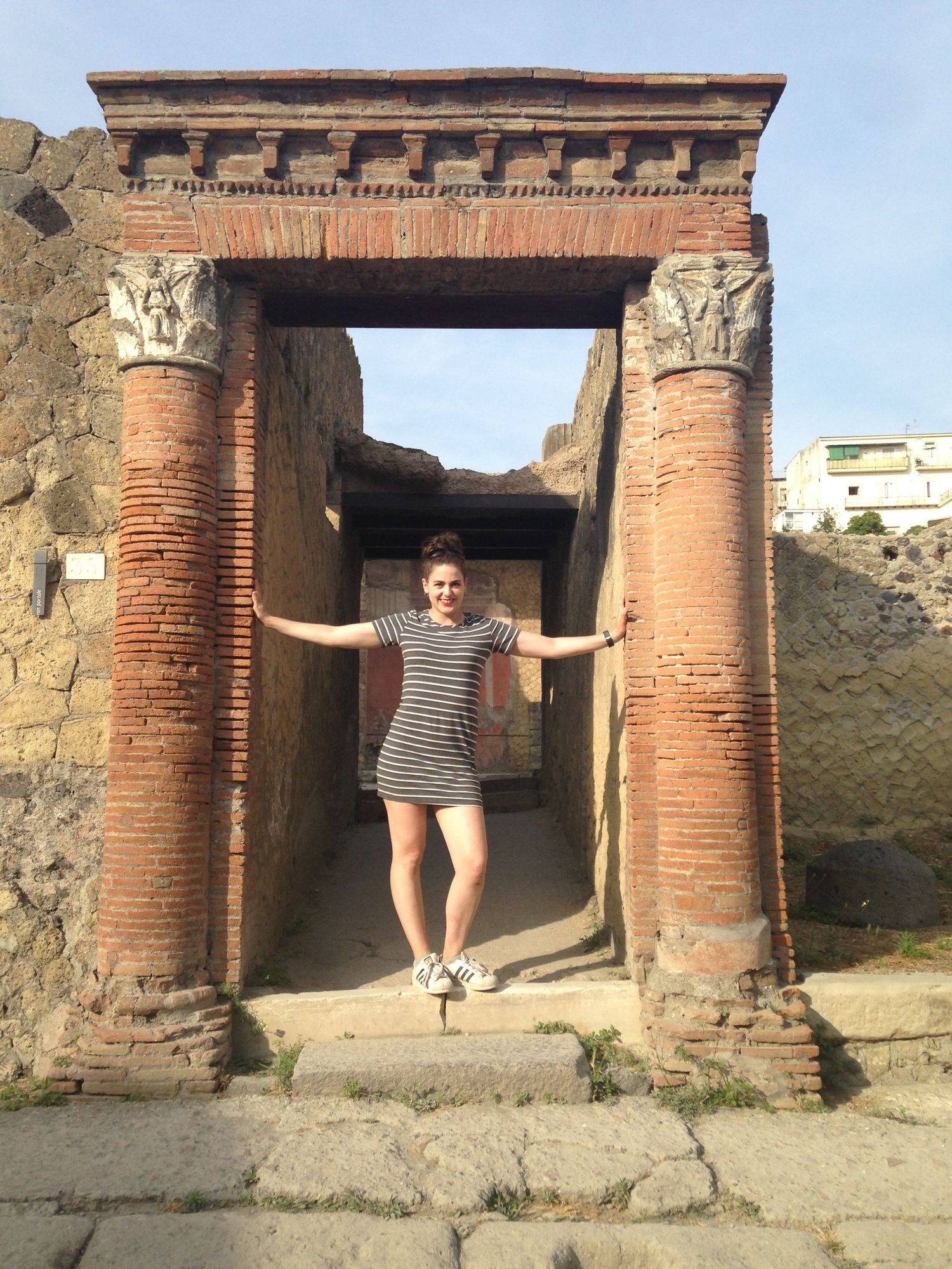 Young woman posing between ancient Roman brick columns at Pompeii ruins, wearing striped shirt and shorts.