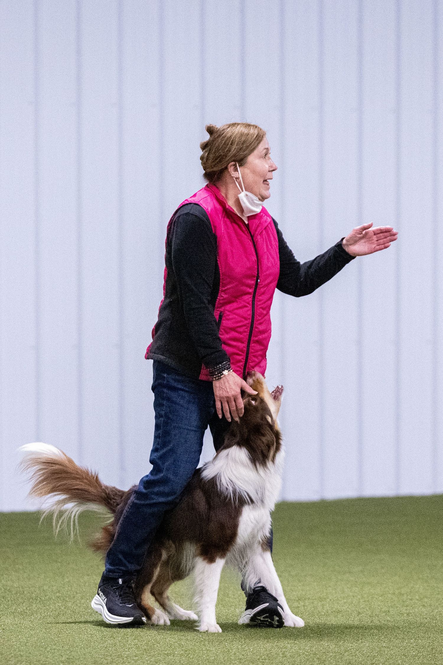 Woman in pink vest directing a Border Collie walking beside her during indoor obedience training.