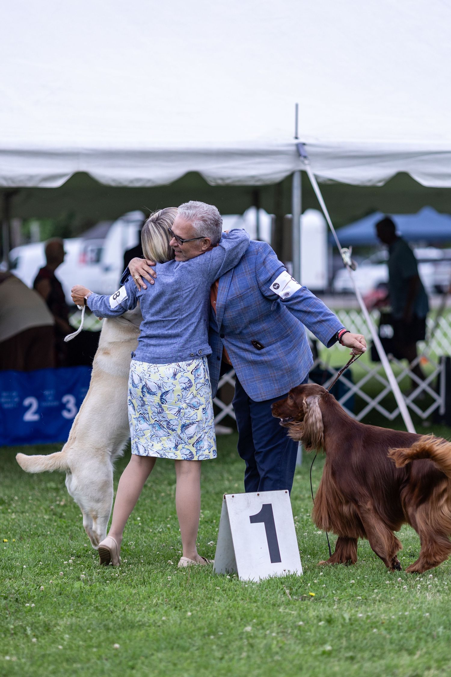 Two dog handlers embrace near a number one marker while their dogs stand nearby at a dog show.