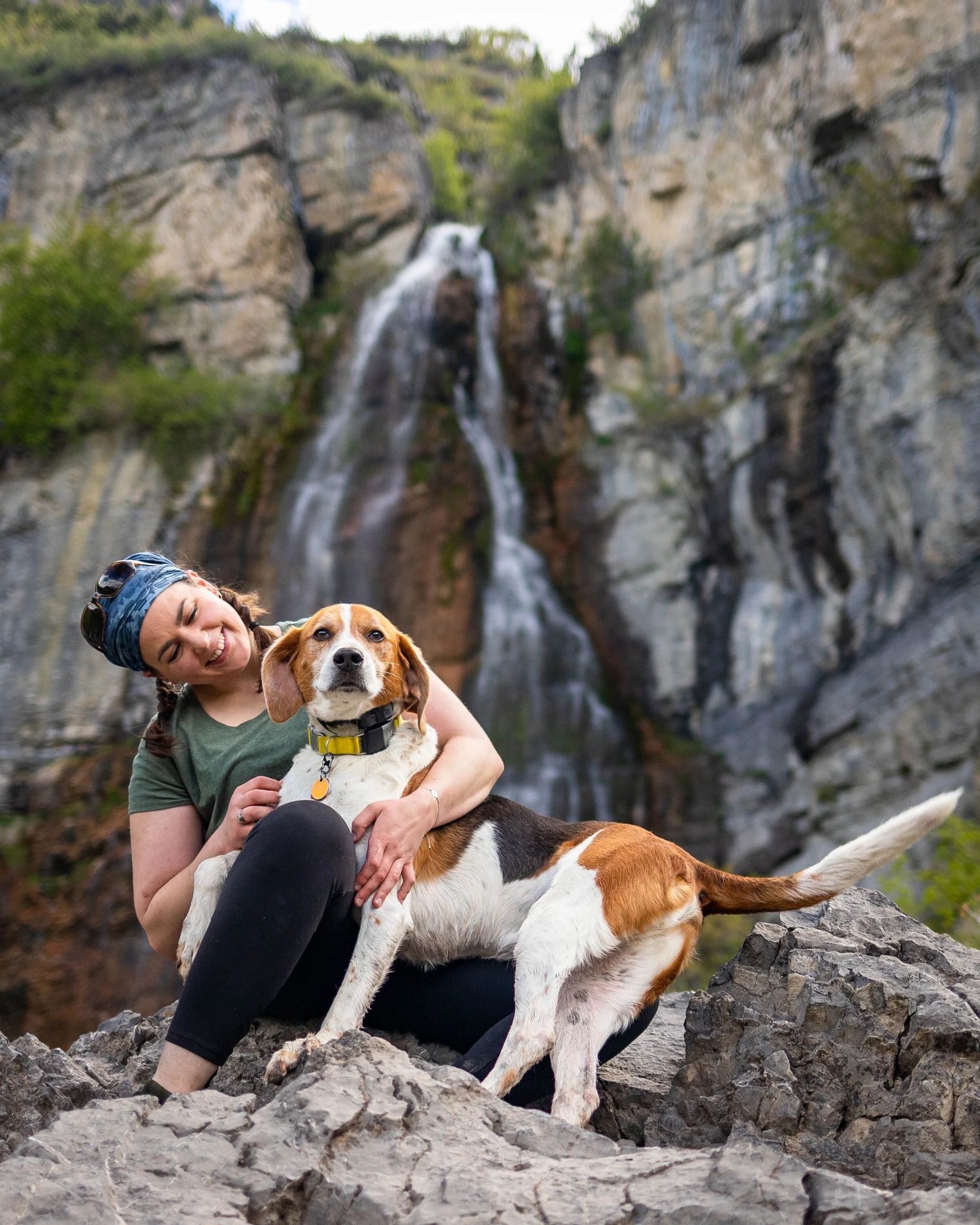Woman hugging a beagle dog on rocks in front of a scenic waterfall cascading down a rocky cliff.