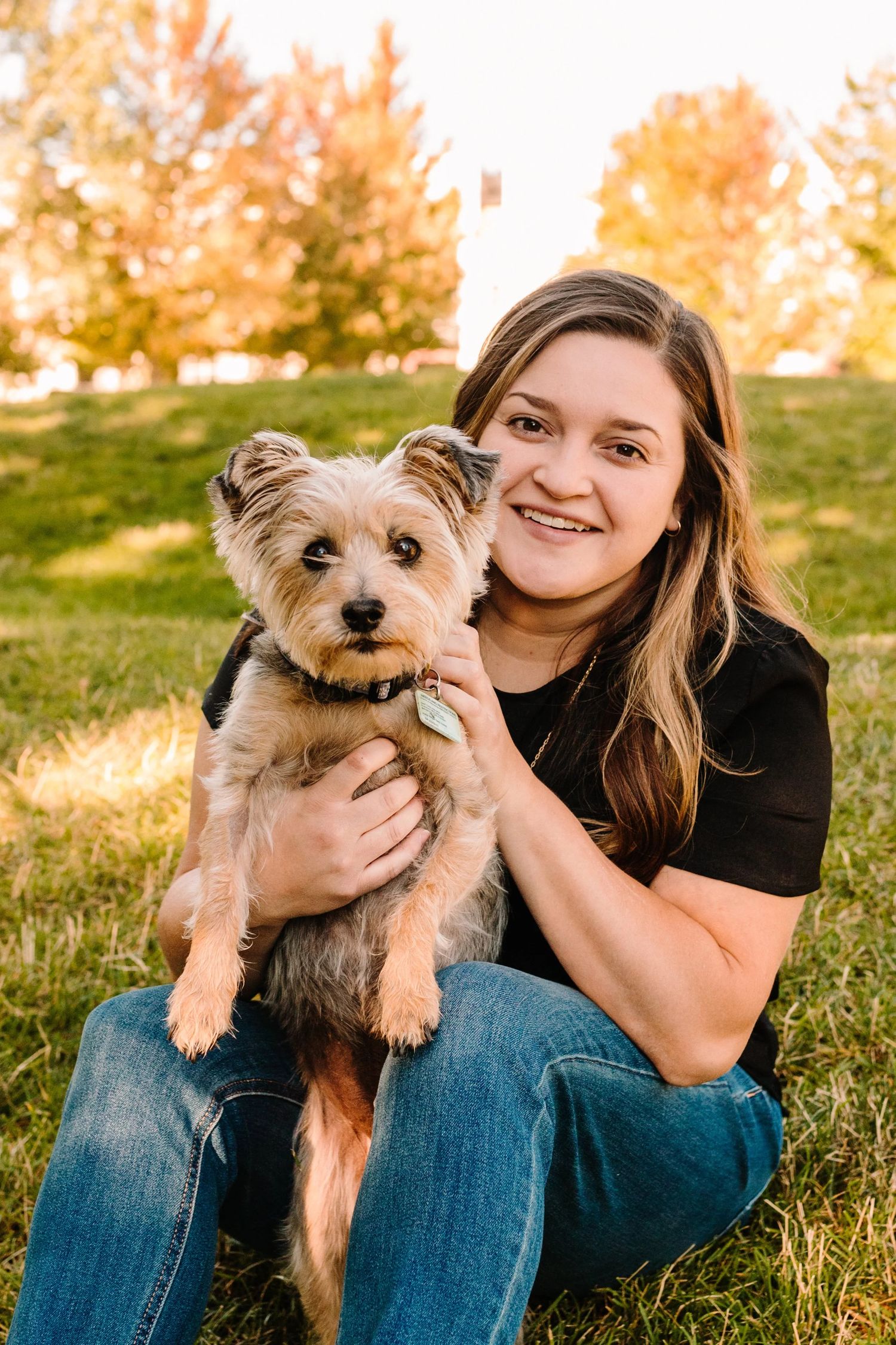 Smiling young woman sitting on grass holding a small Yorkshire Terrier dog outdoors with autumn trees in background.