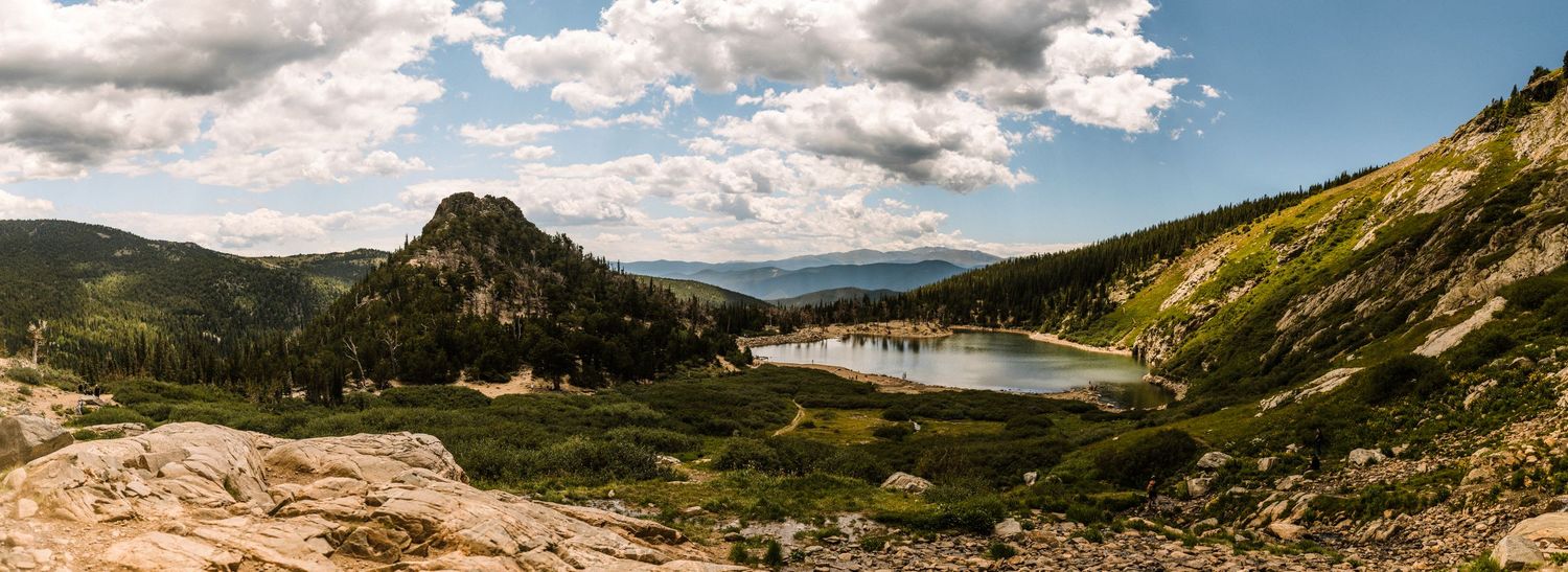 Panoramic view of a serene alpine lake nestled in a mountain valley with rocky peaks, green slopes, and dramatic cloudy skies.