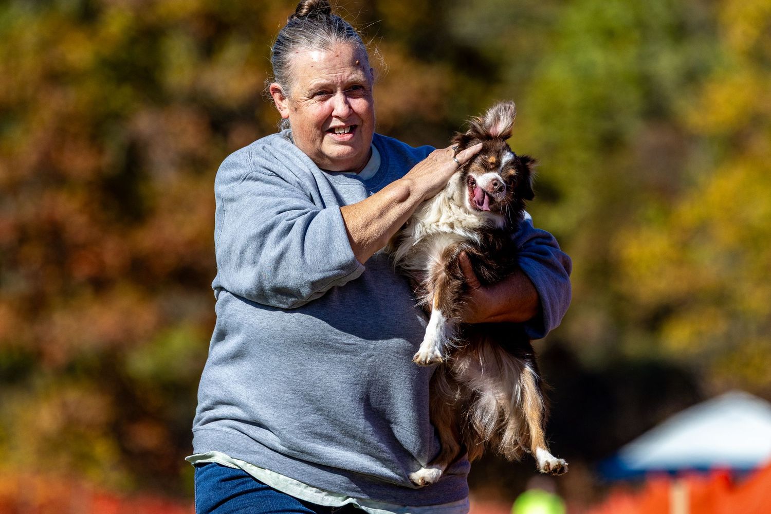 Woman joyfully holding a brown and white Australian Shepherd dog outdoors in autumn park setting.