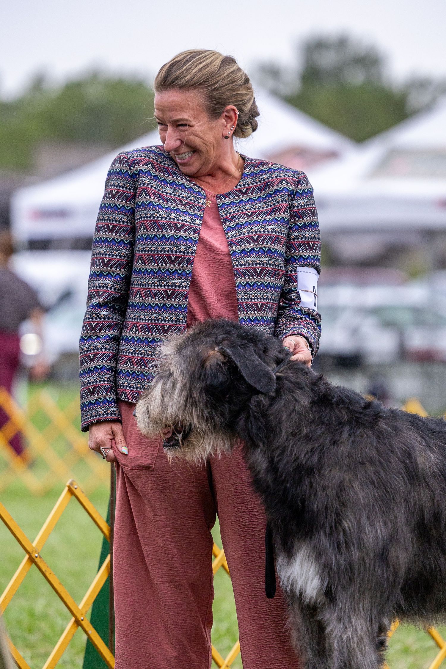 Woman in colorful jacket laughing with large gray Irish Wolfhound at outdoor dog show competition.