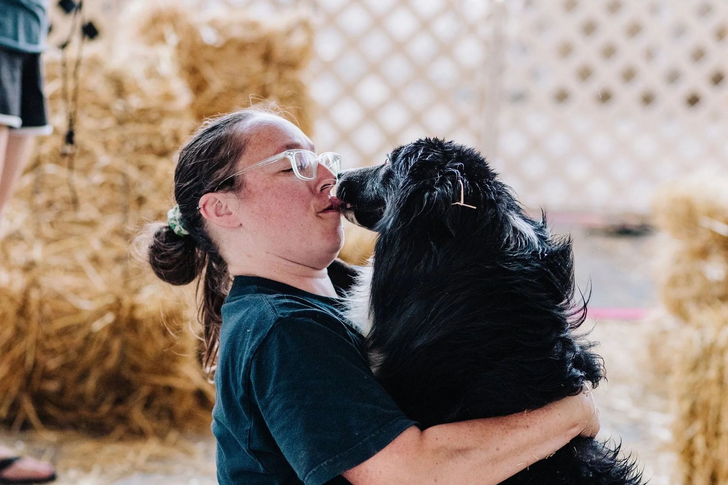 Woman with glasses hugging and kissing a fluffy black dog near hay bales during a Barn Hunt trial.