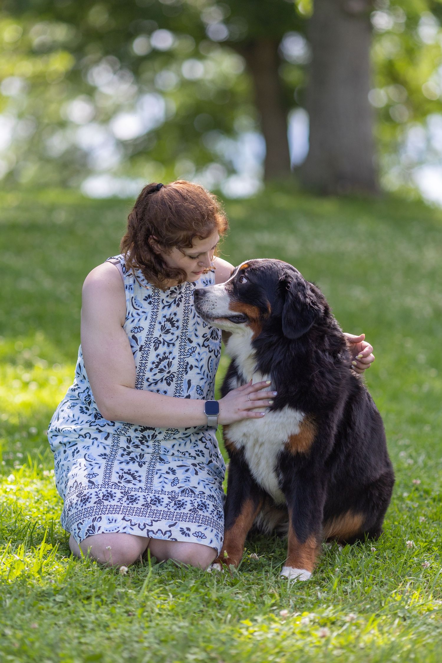 Woman in floral dress kneeling on grass sharing a tender moment with a Bernese Mountain Dog in a park.