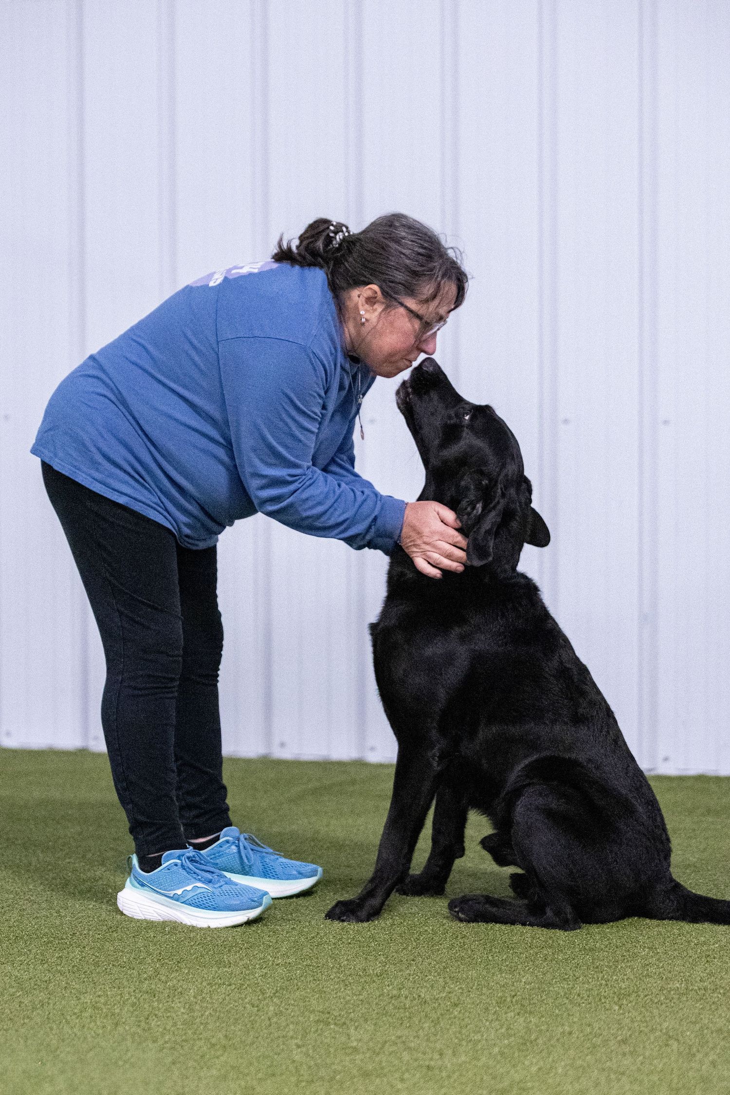 Woman in blue top leaning down as black Labrador reaches up to sniff her face during an indoor agility competition.