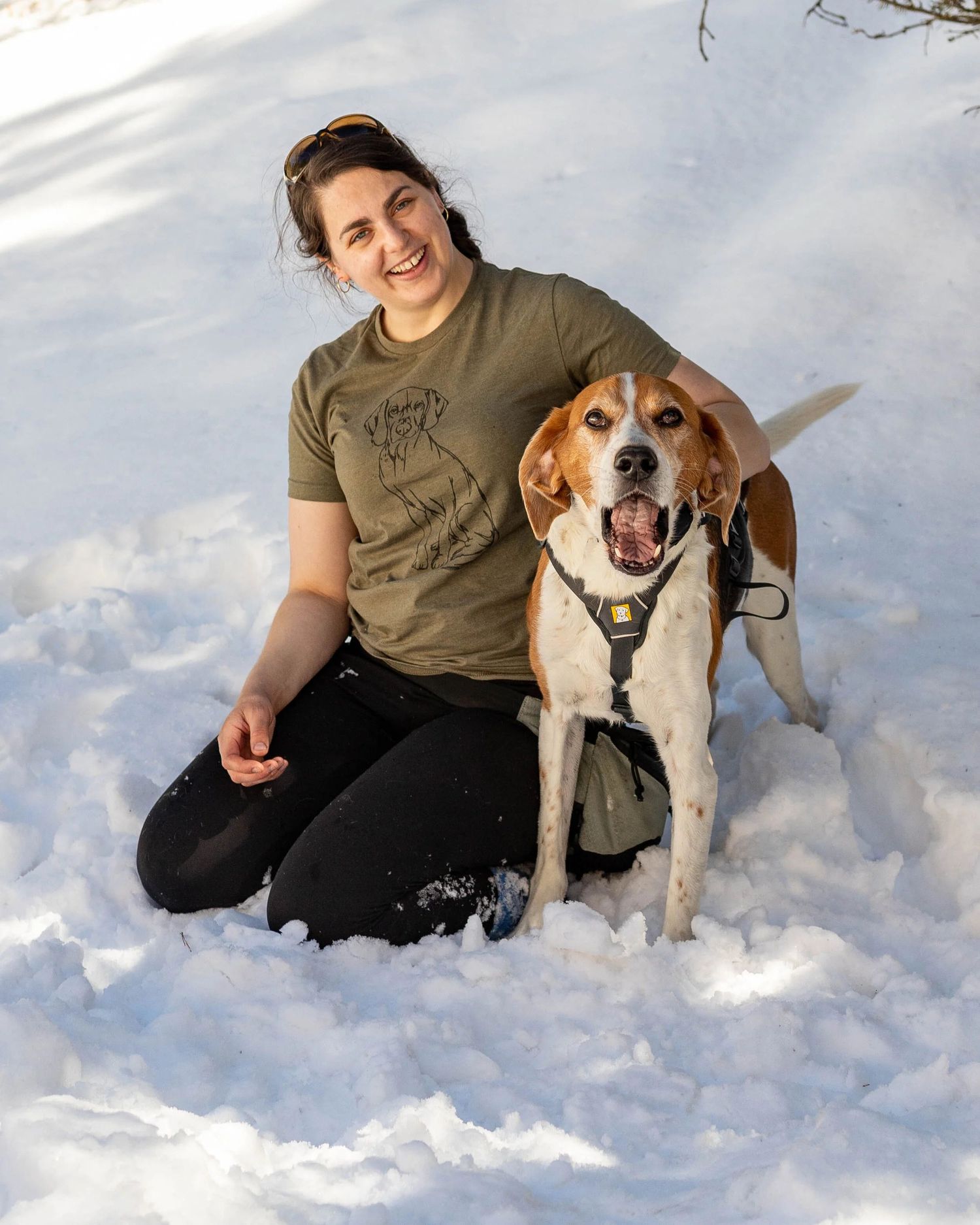 Smiling woman in olive t-shirt sitting in snow with a beagle dog wearing a harness on a sunny winter day.