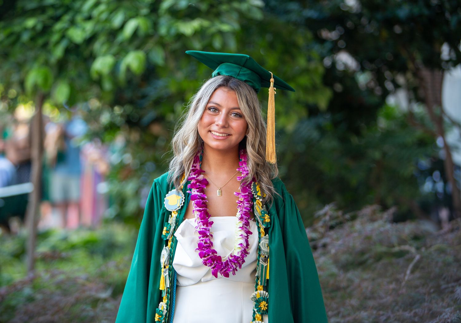 Photograph by Allie of a smiling graduate outdoors, wearing a green cap and gown decorated with colorful honor cords, and medals.