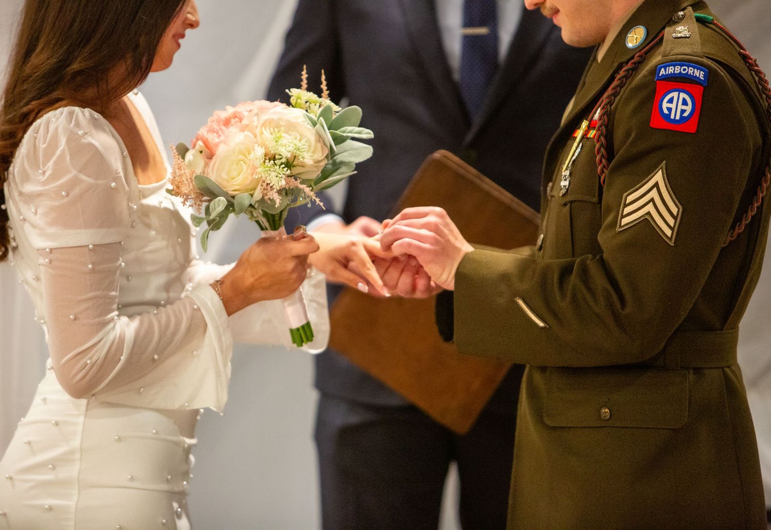 Photograph by Allie capturing a military wedding ceremony as a bride holding a pastel bouquet places a ring on the groom’s finger.