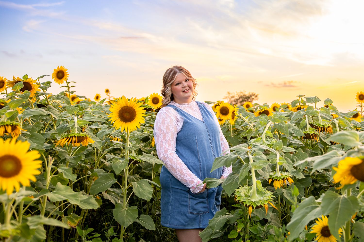 Smiling young woman in blue jumper standing in vibrant sunflower field at golden sunset.