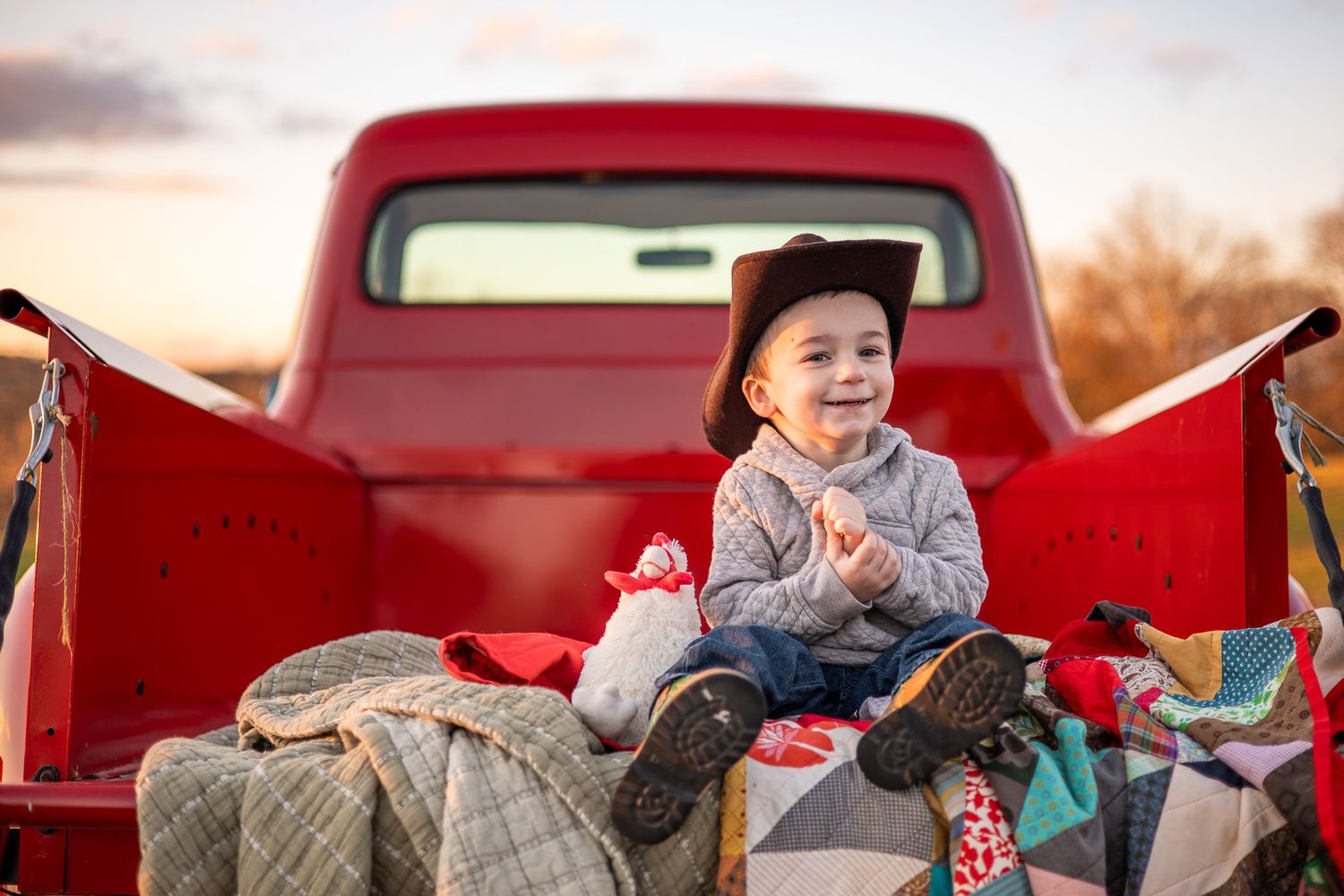 Adorable toddler in cowboy hat sitting in red truck bed with quilts at golden sunset.