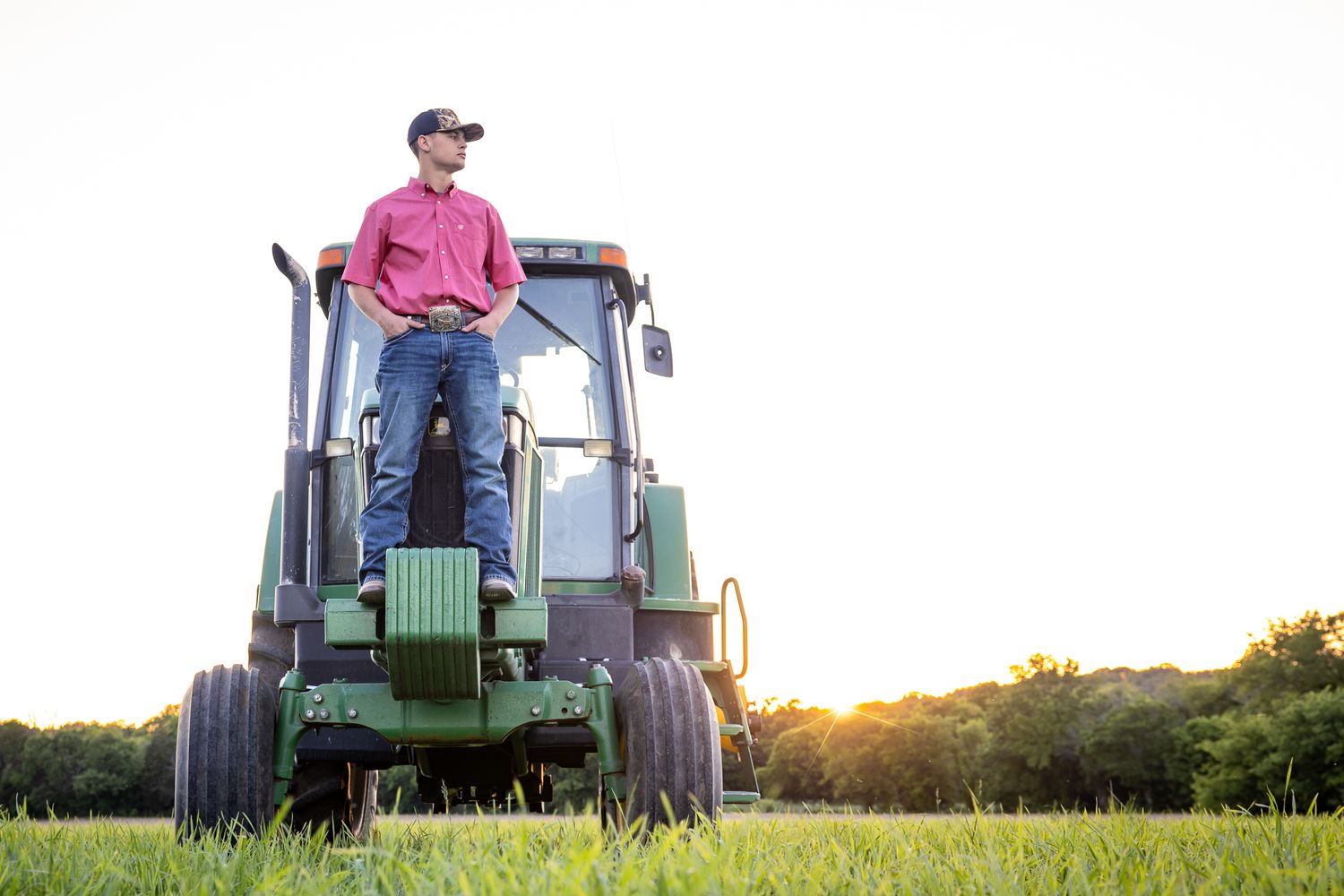 Young man in red shirt and cap standing on green John Deere tractor in field at golden sunset.