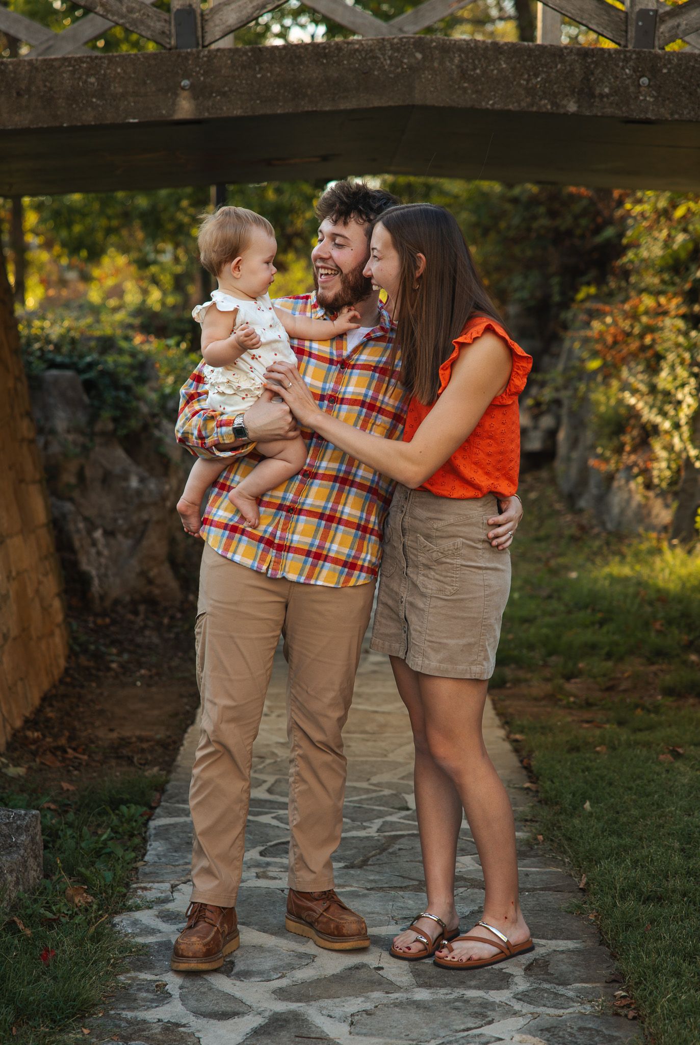 Young couple with baby standing under wooden arbor in lush garden, family portrait photography.
