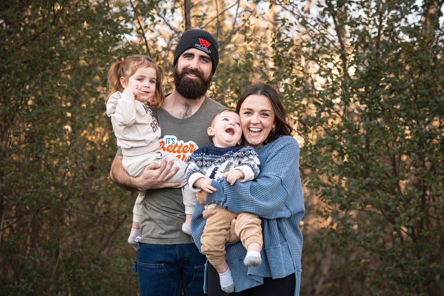 Happy young family with bearded dad and mom holding two small children in autumn woods.