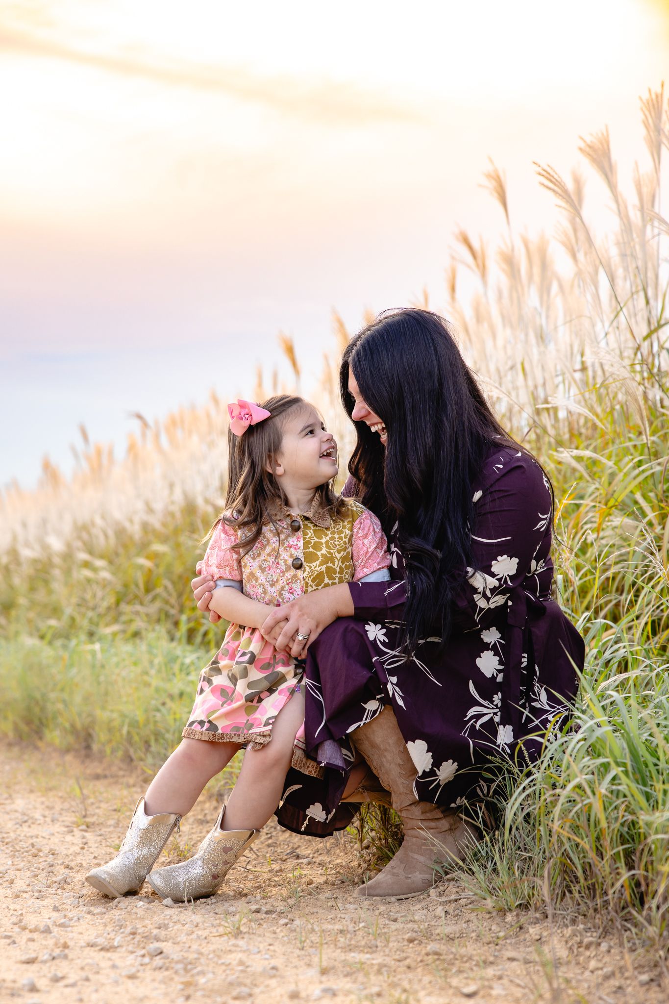 A mother and young daughter share a joyful moment outdoors in floral dresses, surrounded by tall golden grasses at sunset.
