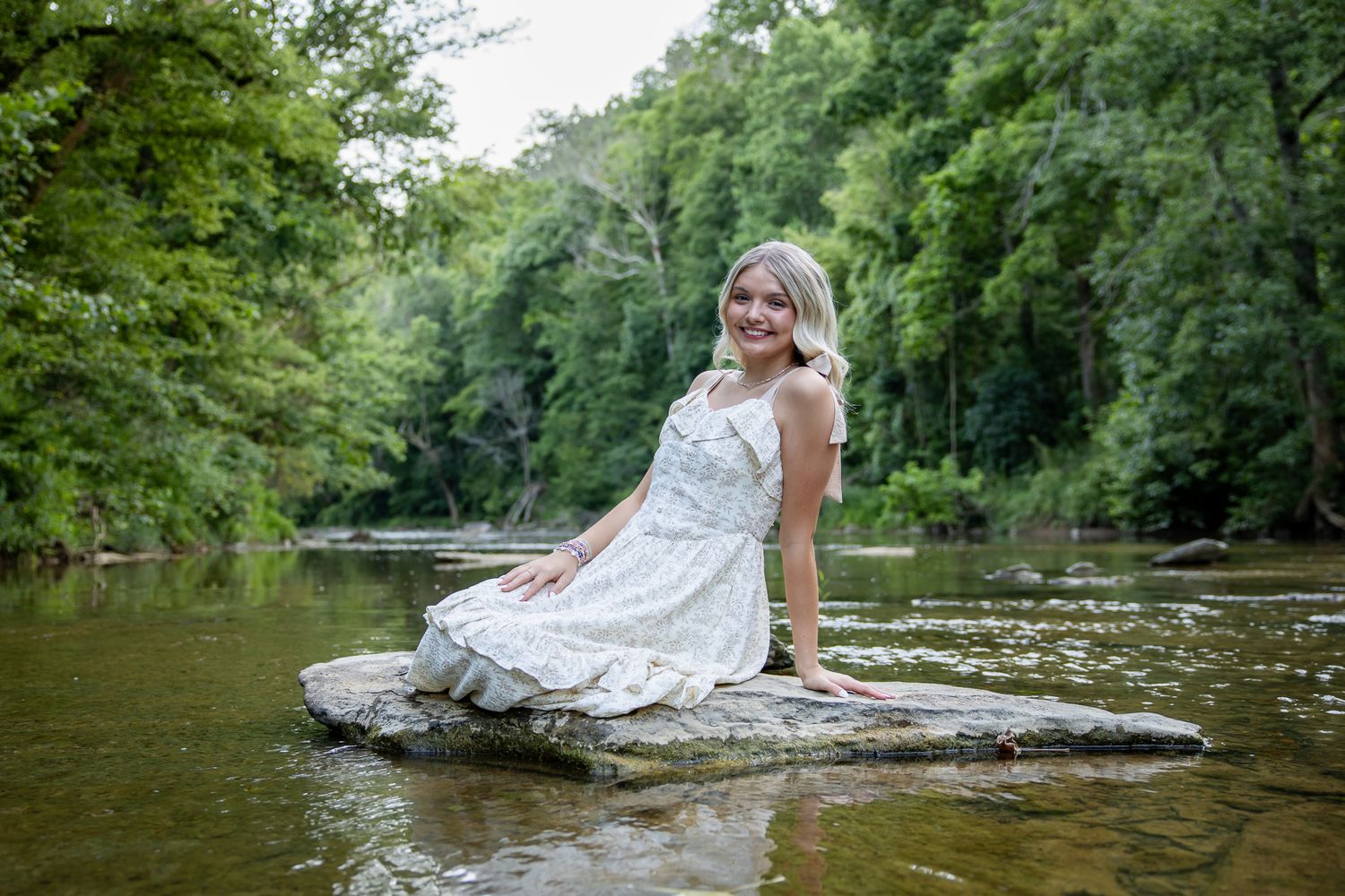 Smiling young woman in white floral dress sitting on rocks by a serene forest river.