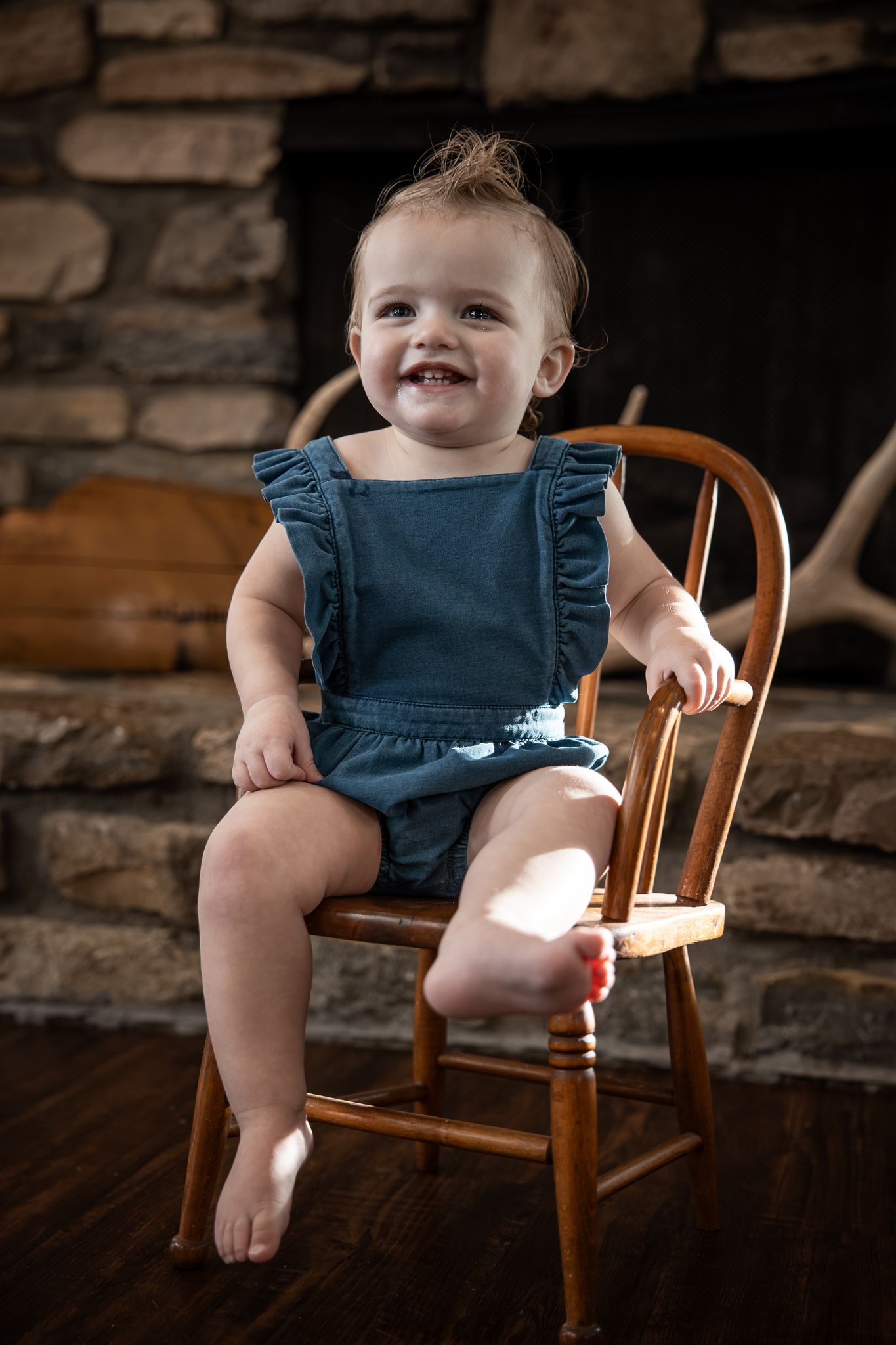Adorable smiling baby girl in blue romper sitting on wooden chair by stone fireplace.
