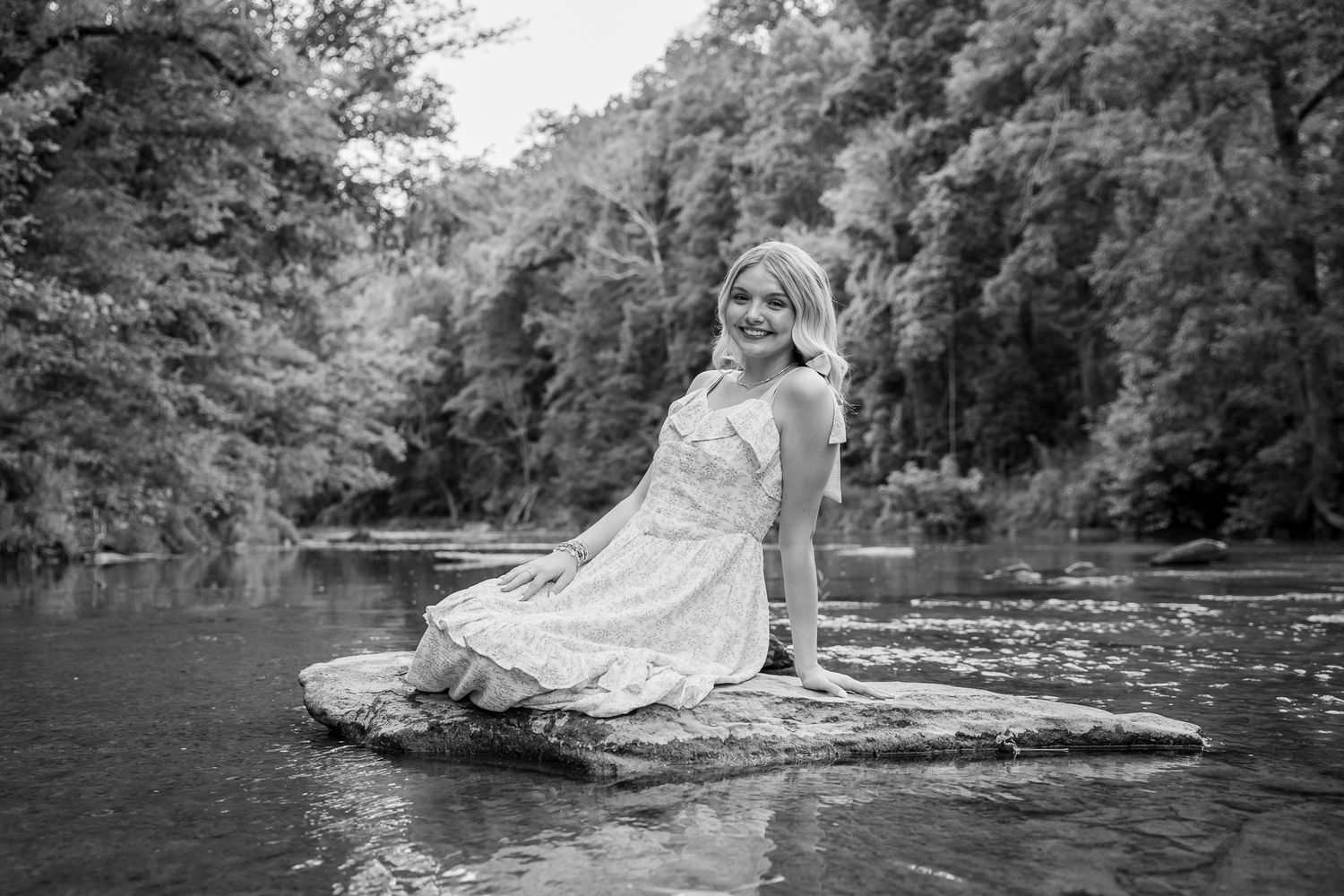 Black and white portrait of a smiling woman in a floral dress sitting on rocks by a river.