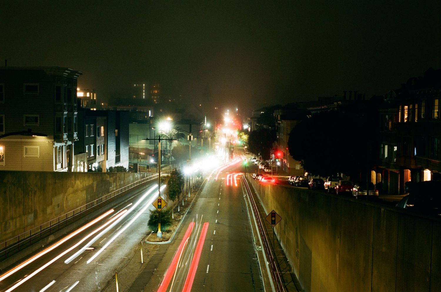 A nighttime long-exposure shot shows light trails from traffic moving along a city street flanked by retaining walls and buildings.