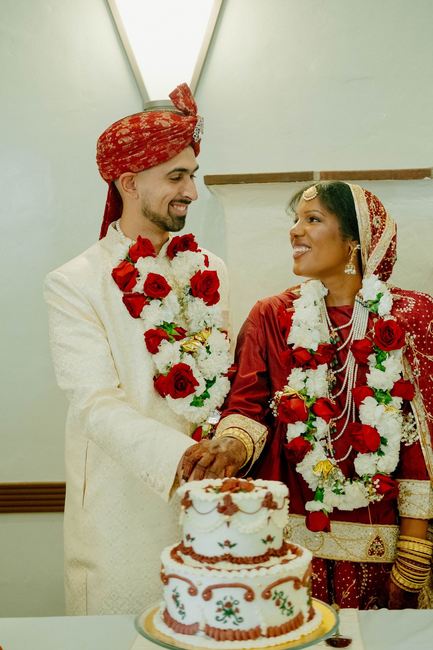 The couple smiles at each other while cutting their tiered, decorated wedding cake together.