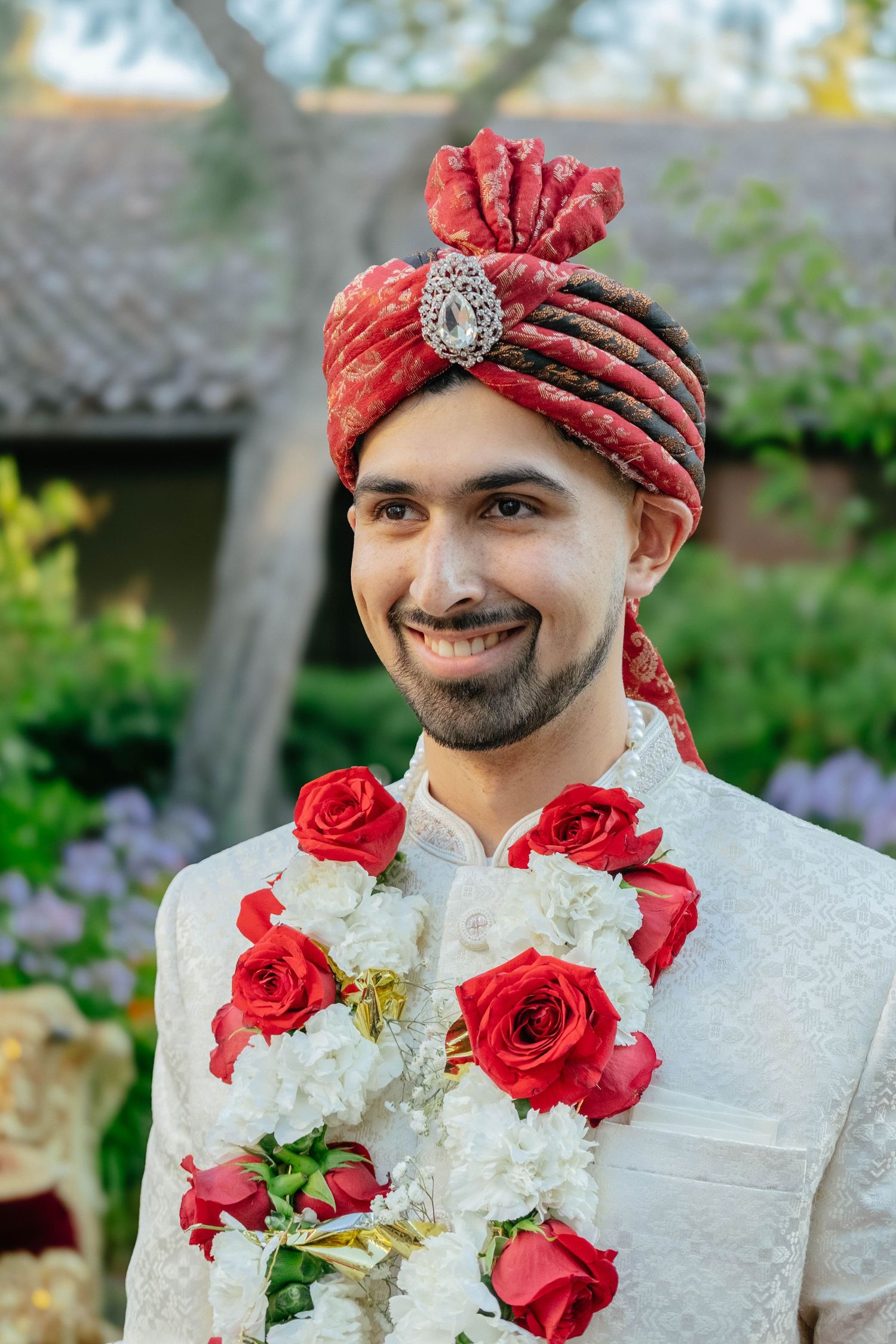 A portrait focuses on the groom wearing a traditional red turban and a garland of red and white roses.