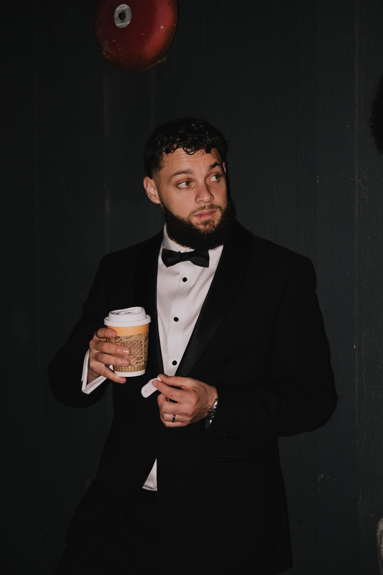 A man in a formal tuxedo holds a coffee cup in a dimly lit setting.