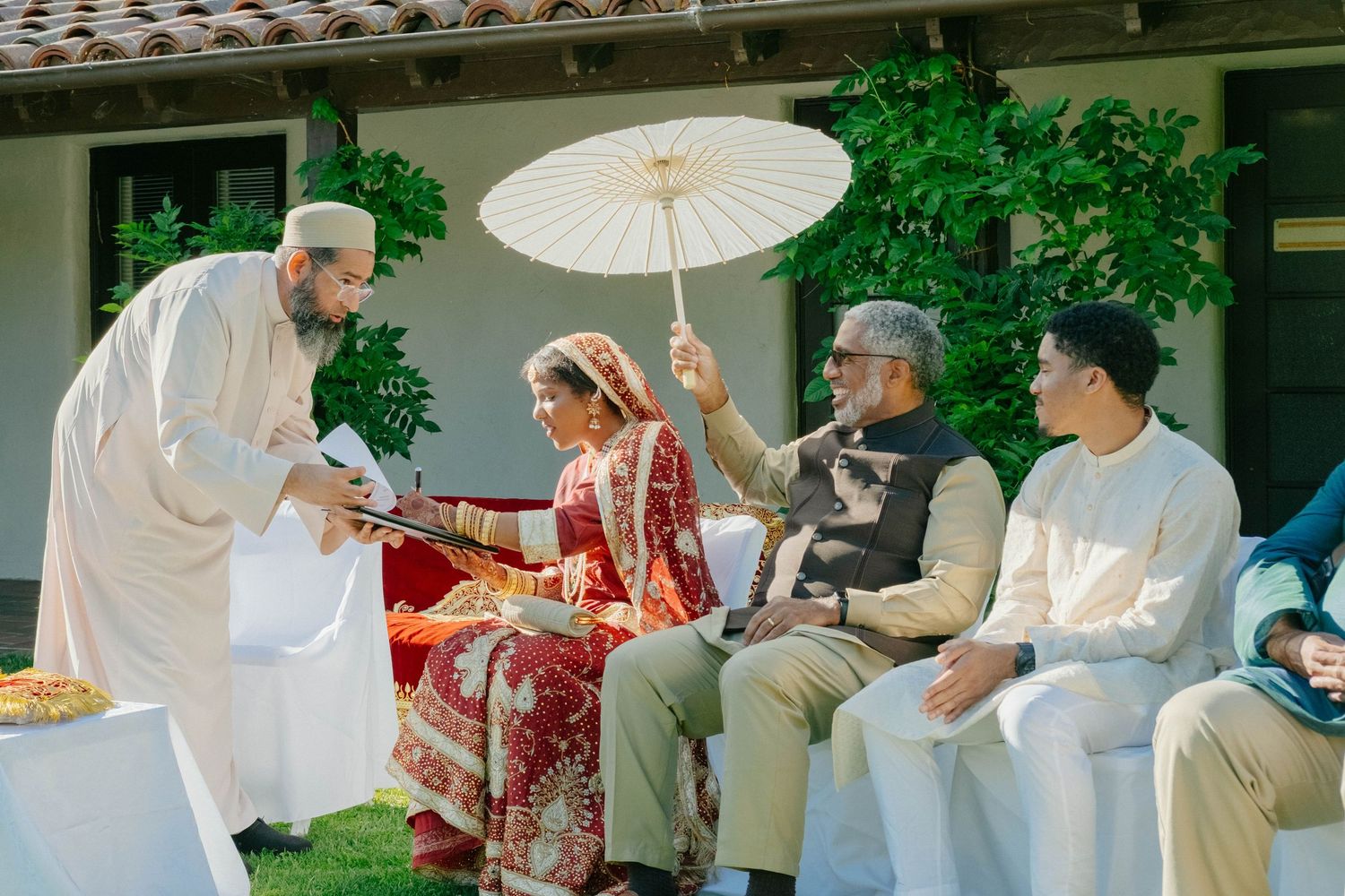 A guest holds a parasol over the bride while a religious official facilitates the ceremony.