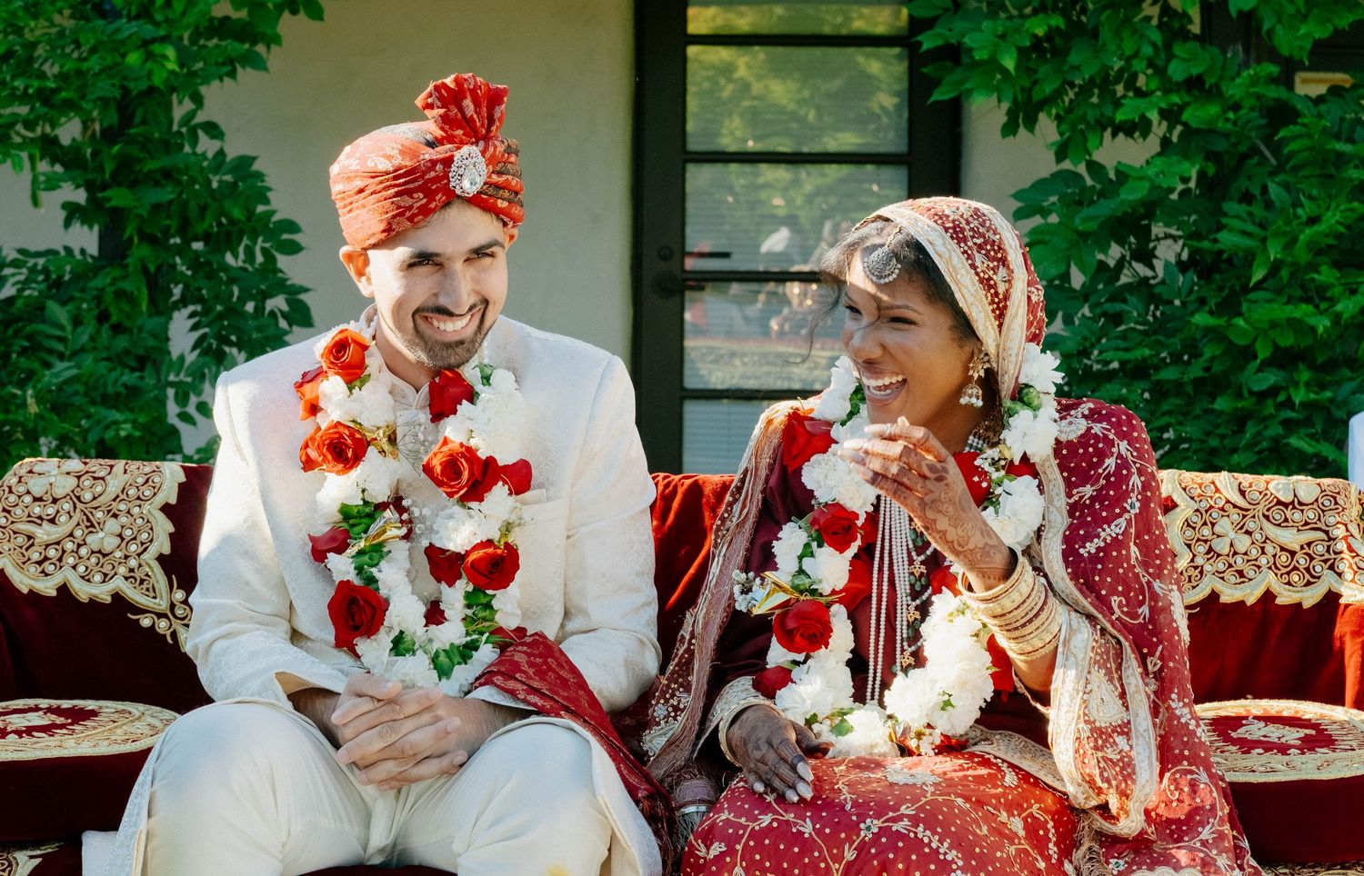The bride and groom laugh together while sitting on a decorated couch during their outdoor wedding ceremony.