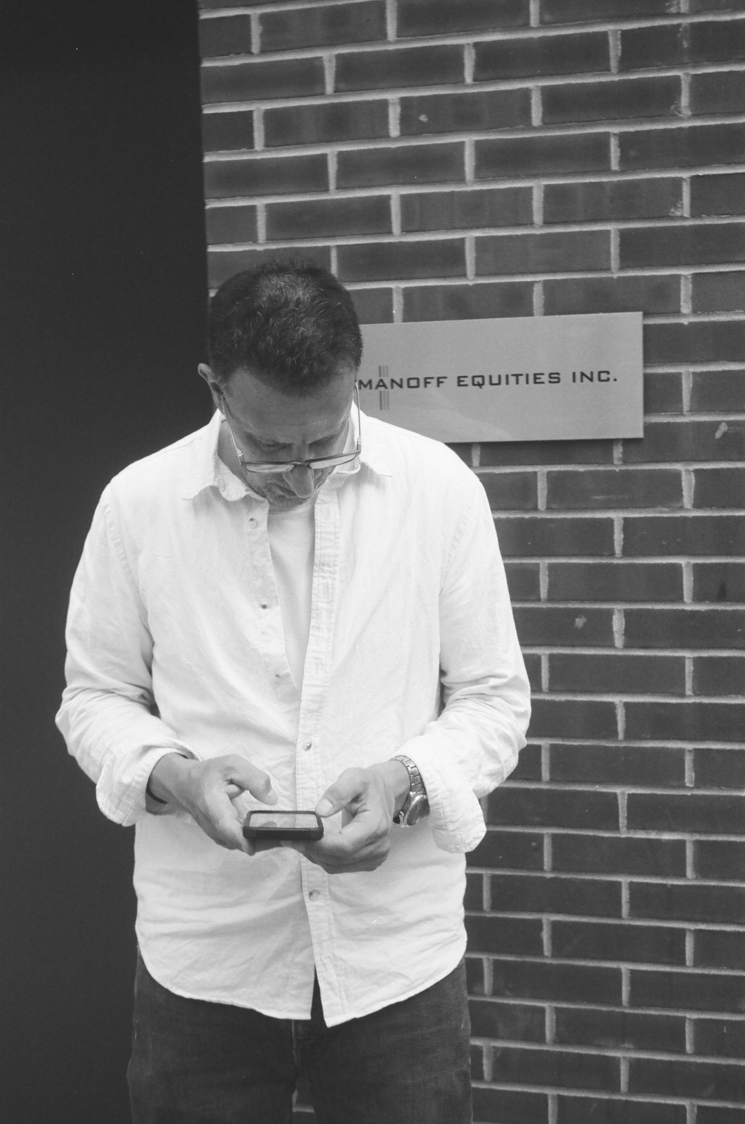 A man in a white shirt checks his phone near brickwork.
