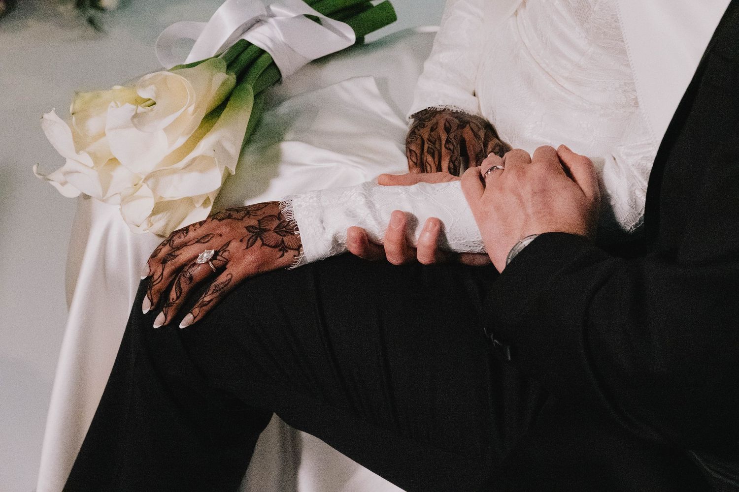 The bride and groom hold hands, displaying henna designs and their wedding rings near a floral arrangement.