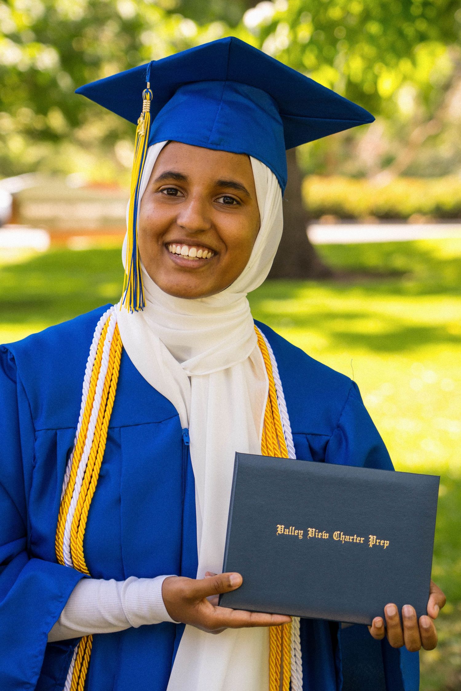 A smiling graduate wears a blue cap and gown while holding diploma.