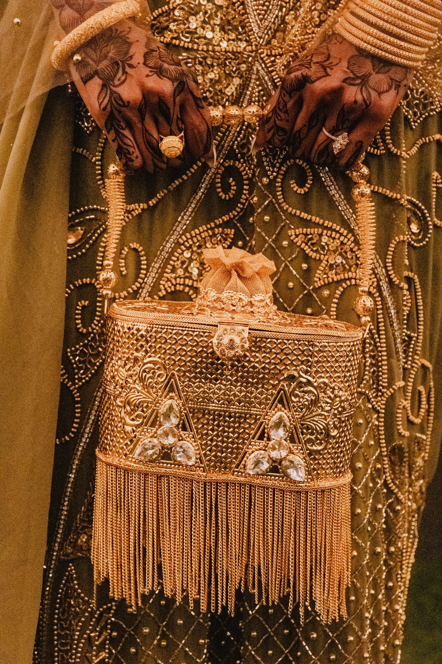 A close-up shot highlights a bride's henna-patterned hands holding an ornate, gold-fringed clutch purse.