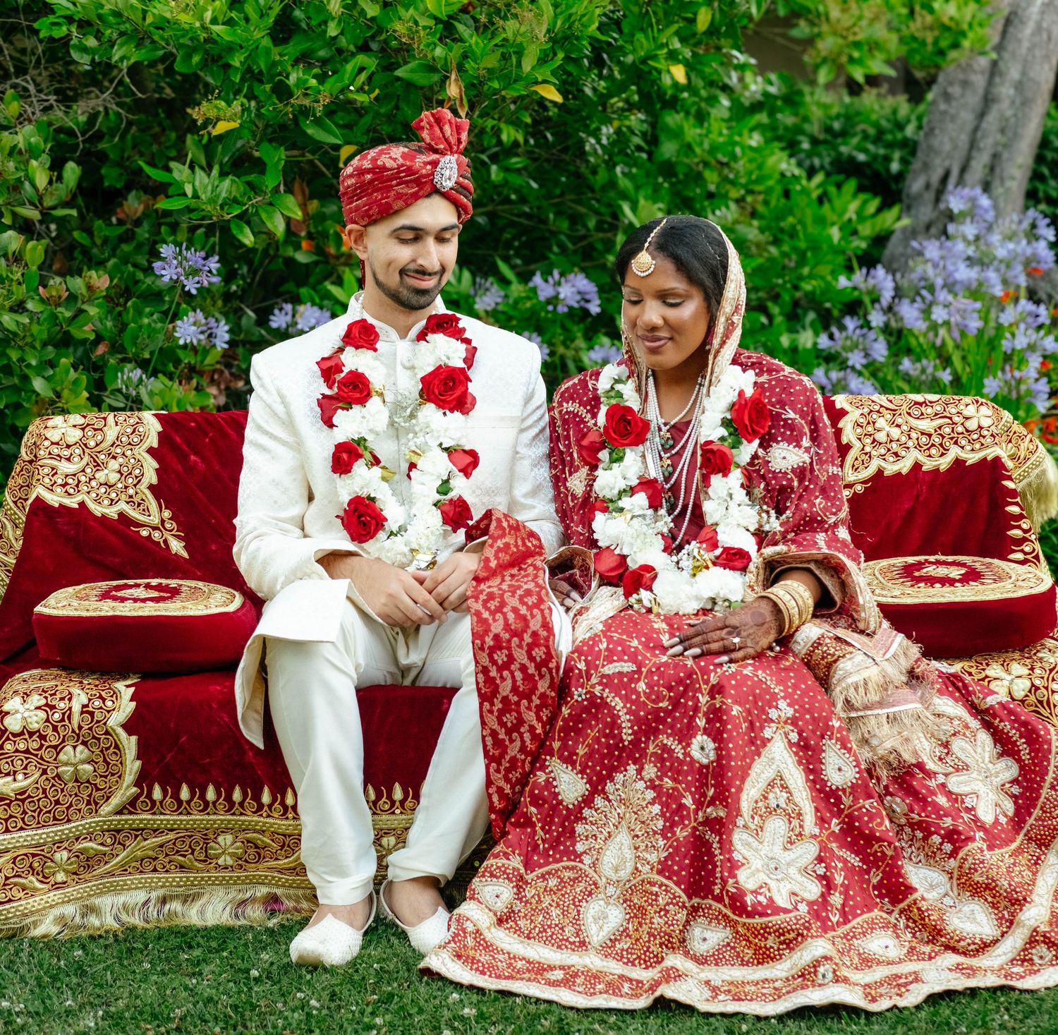 A bride and groom in traditional South Asian attire sit together on a plush, ornate couch.