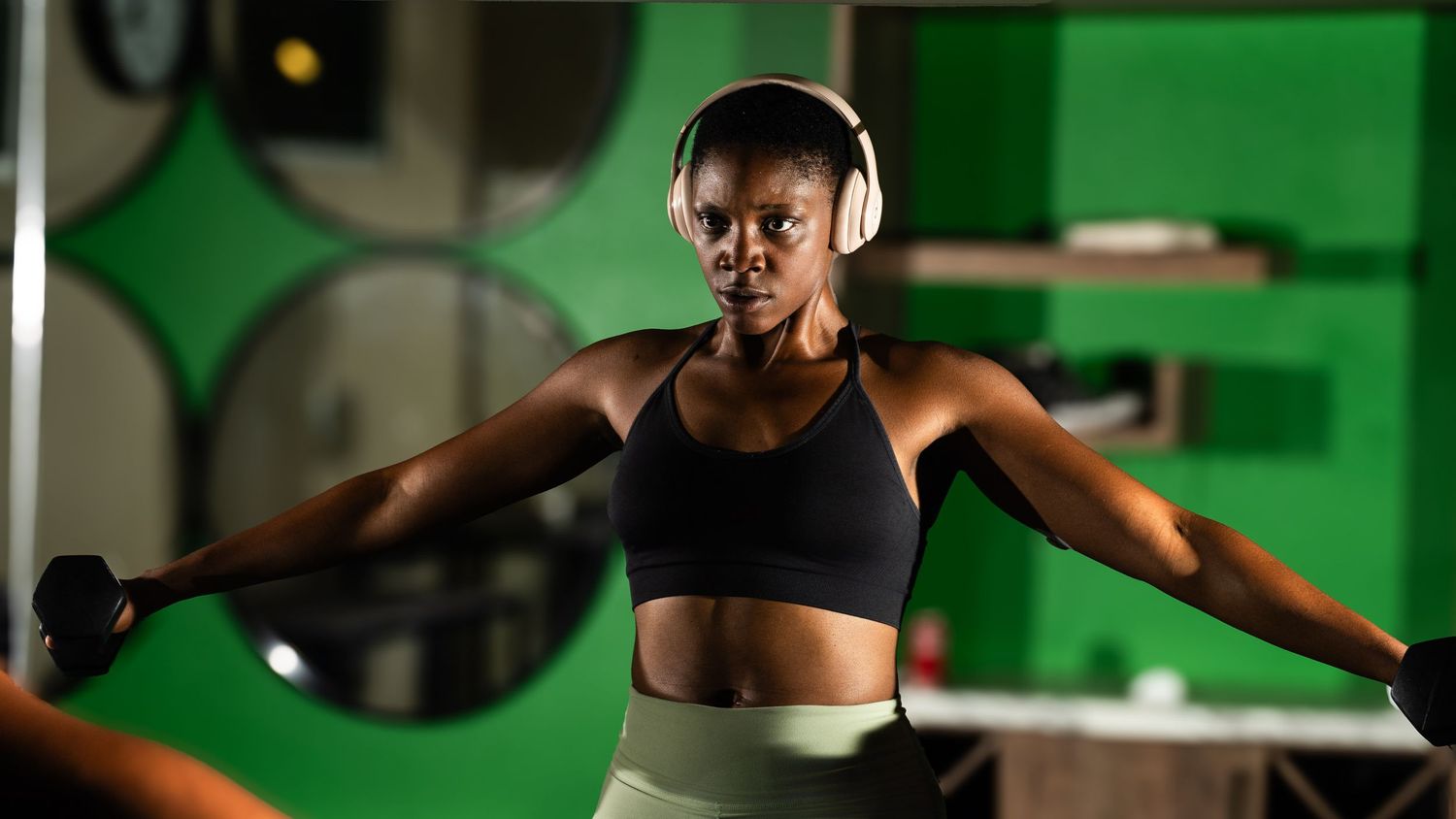 Athletic woman working out with dumbbells wearing headphones in a green lit gym.