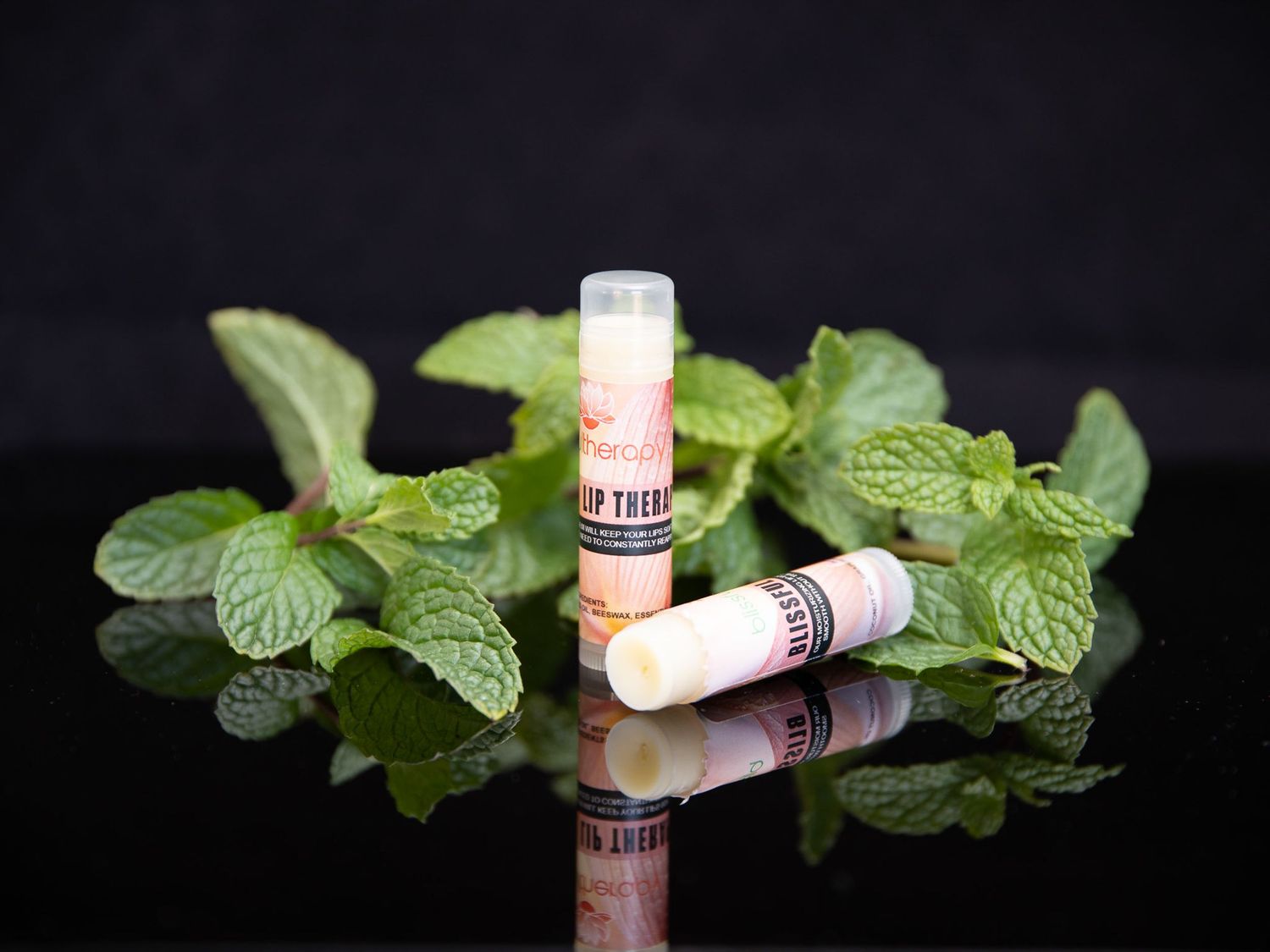 Natural lip-care products arranged with fresh mint leaves on a reflective black surface.