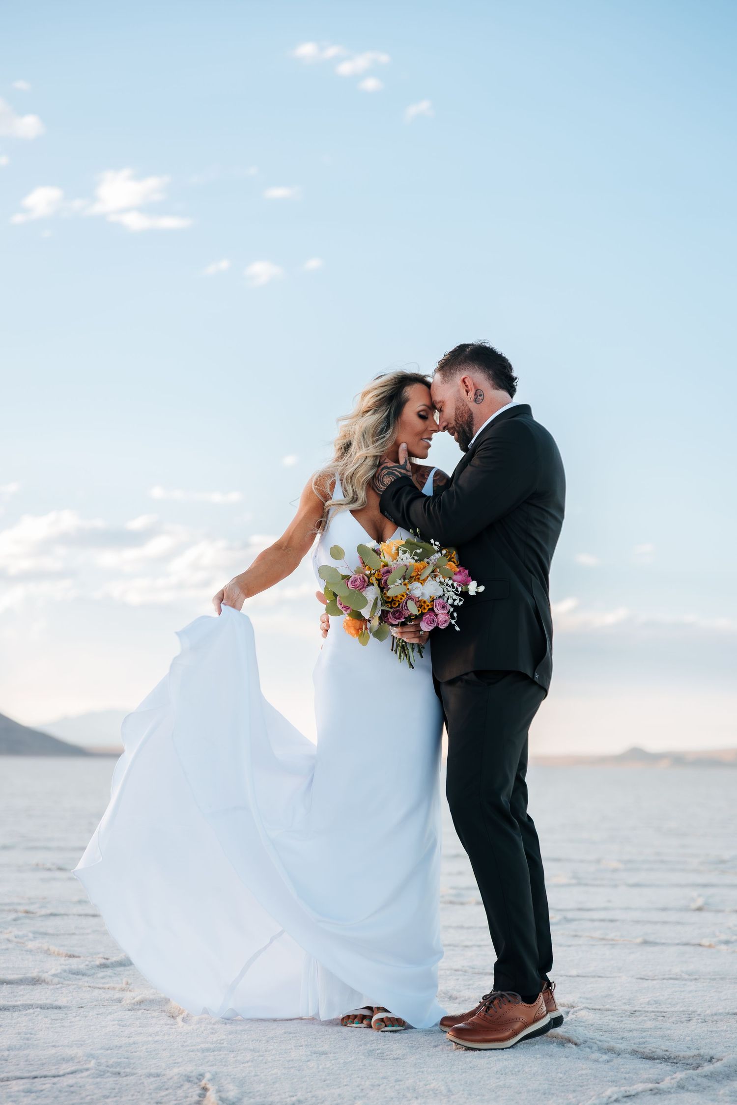 Bride and groom share a romantic moment on Utah salt flats, her white gown flowing, holding a colorful bouquet. Elopement destinations