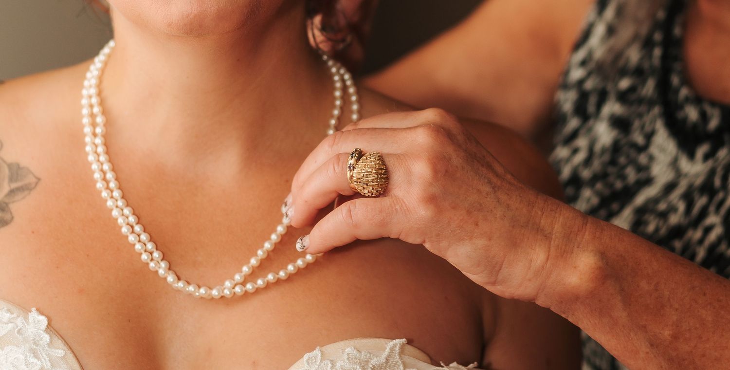 Close-up of a bride wearing a double-strand pearl necklace being fastened by a helper wearing a gold ring.