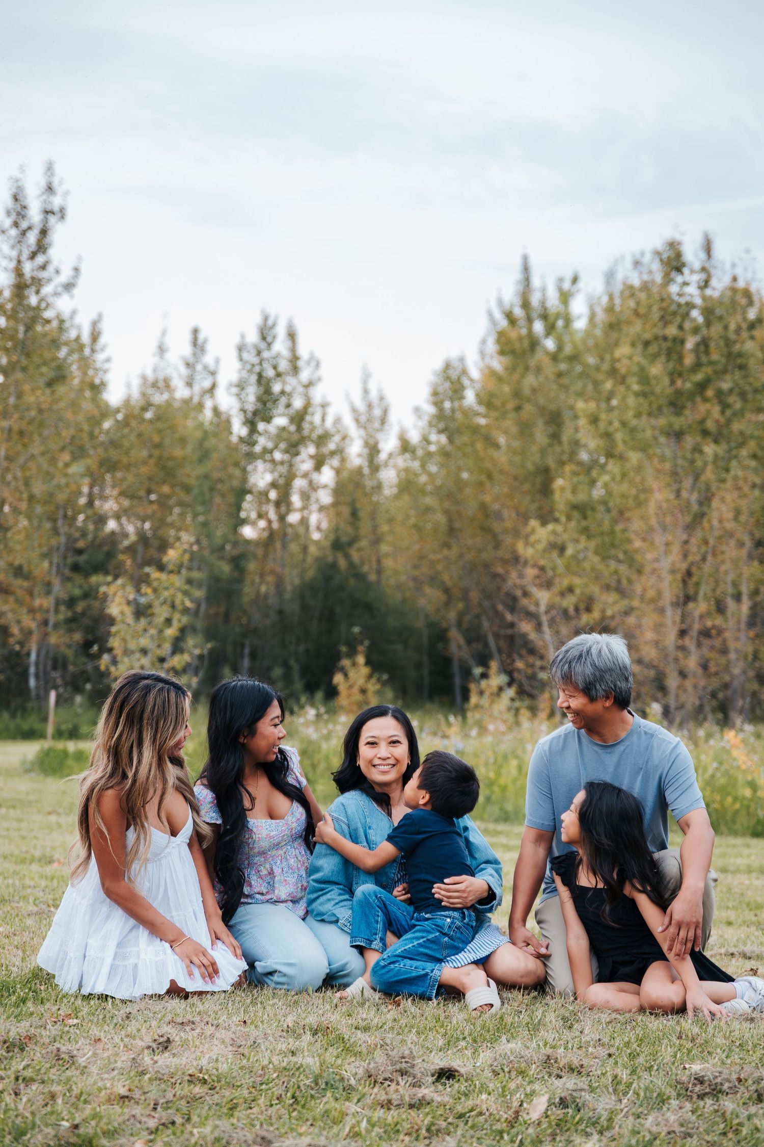 A multigenerational Asian family sitting together on grass in a park, smiling and interacting with a young toddler outdoors. family photos