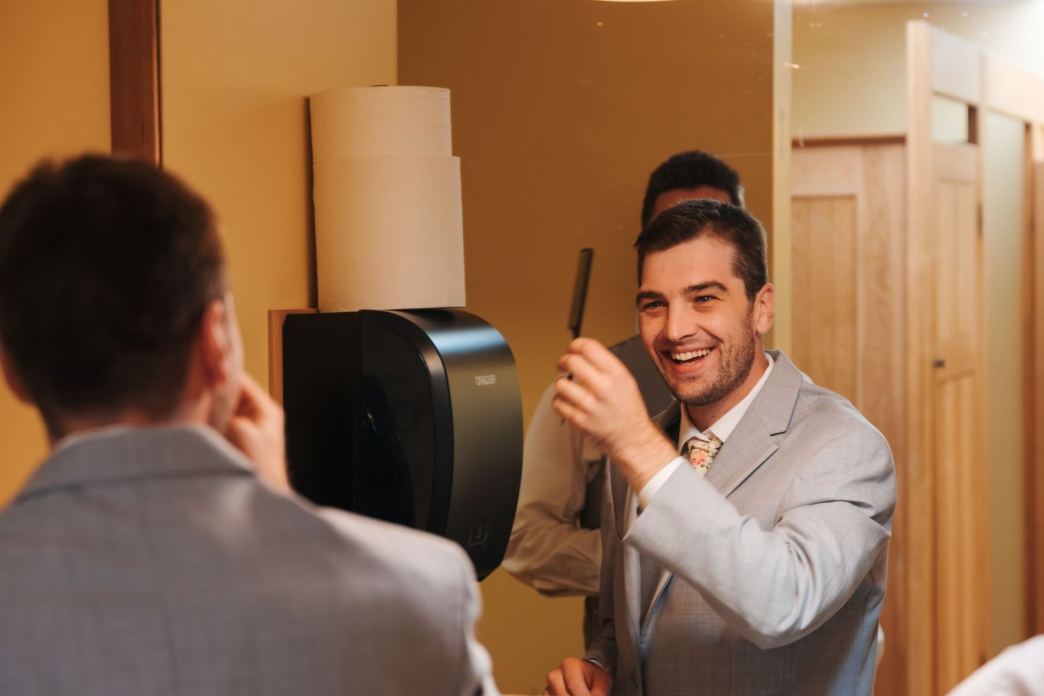 Smiling man in gray suit holds a comb while looking at his reflection in a bathroom mirror.