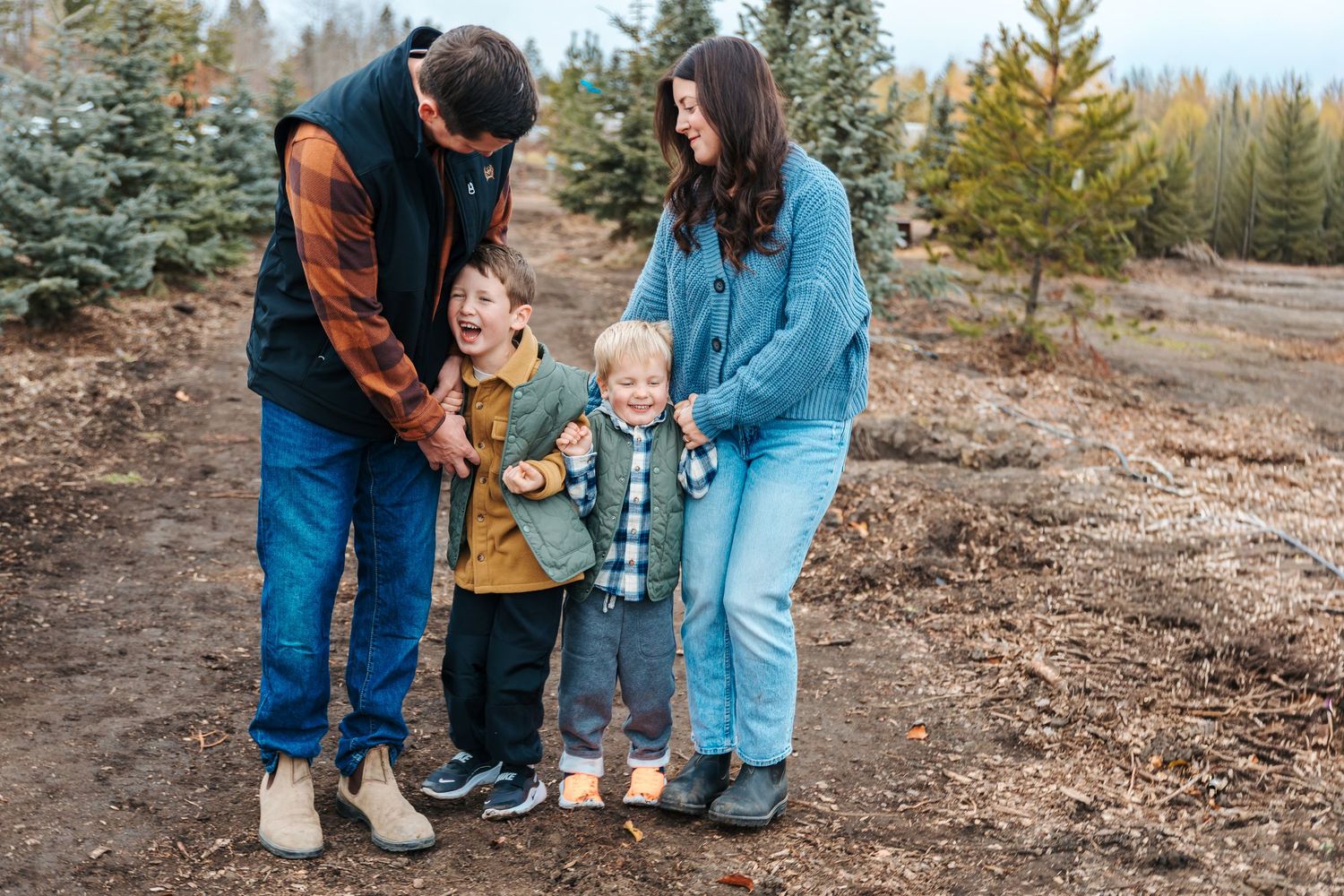 A happy family with two young boys laughing together outdoors at a Christmas tree farm in fall.