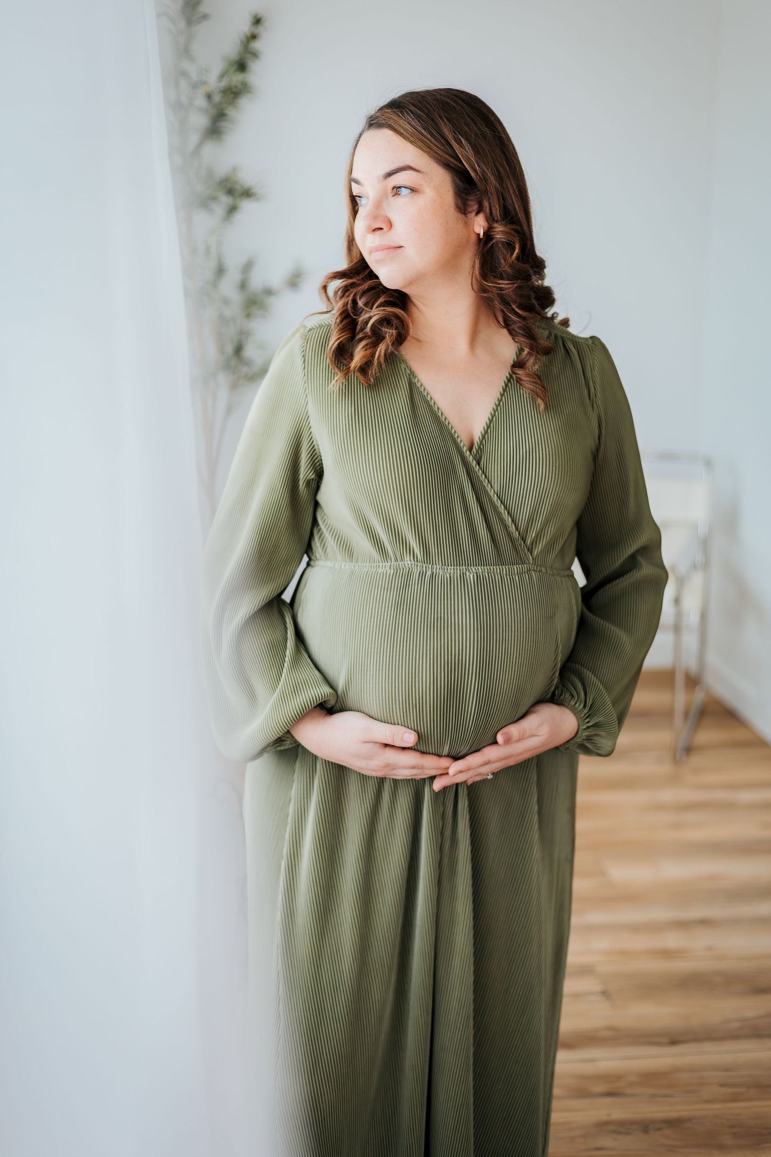 maternity photos at a studio in St.Albert. mom in long green dress holding pregnant belly by window. Family and newborn photographer
