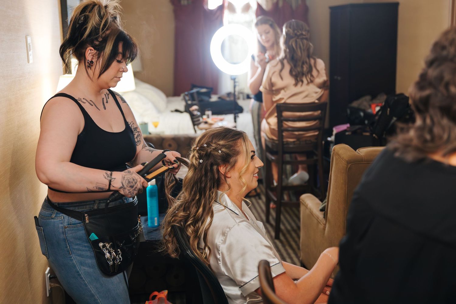 Hair stylist curling a bride's hair in a dressing room with a lighted mirror in the background.