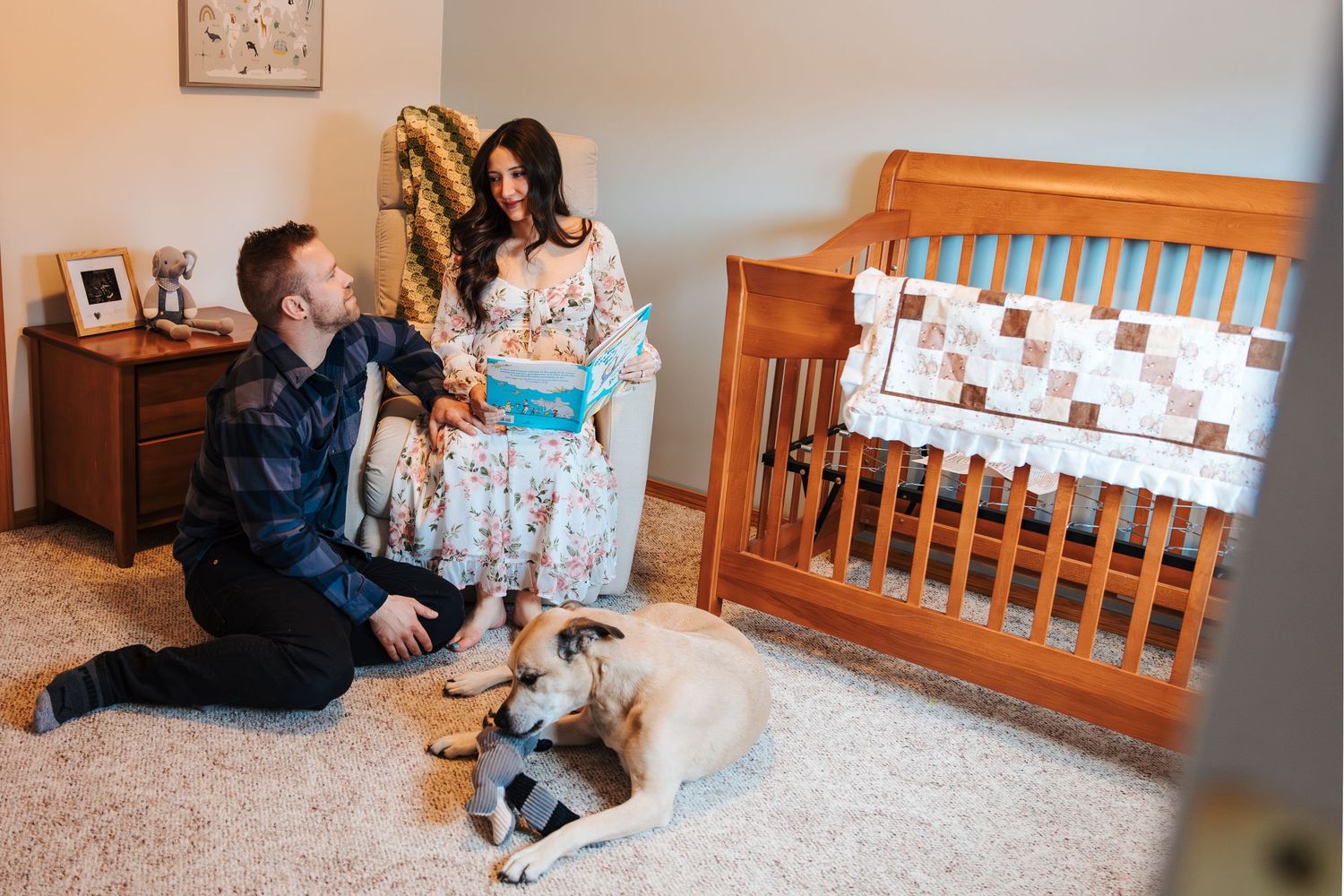 Couple with dog in nursery room next to wooden crib, man kneeling on floor while pregnant woman stands holding item.
