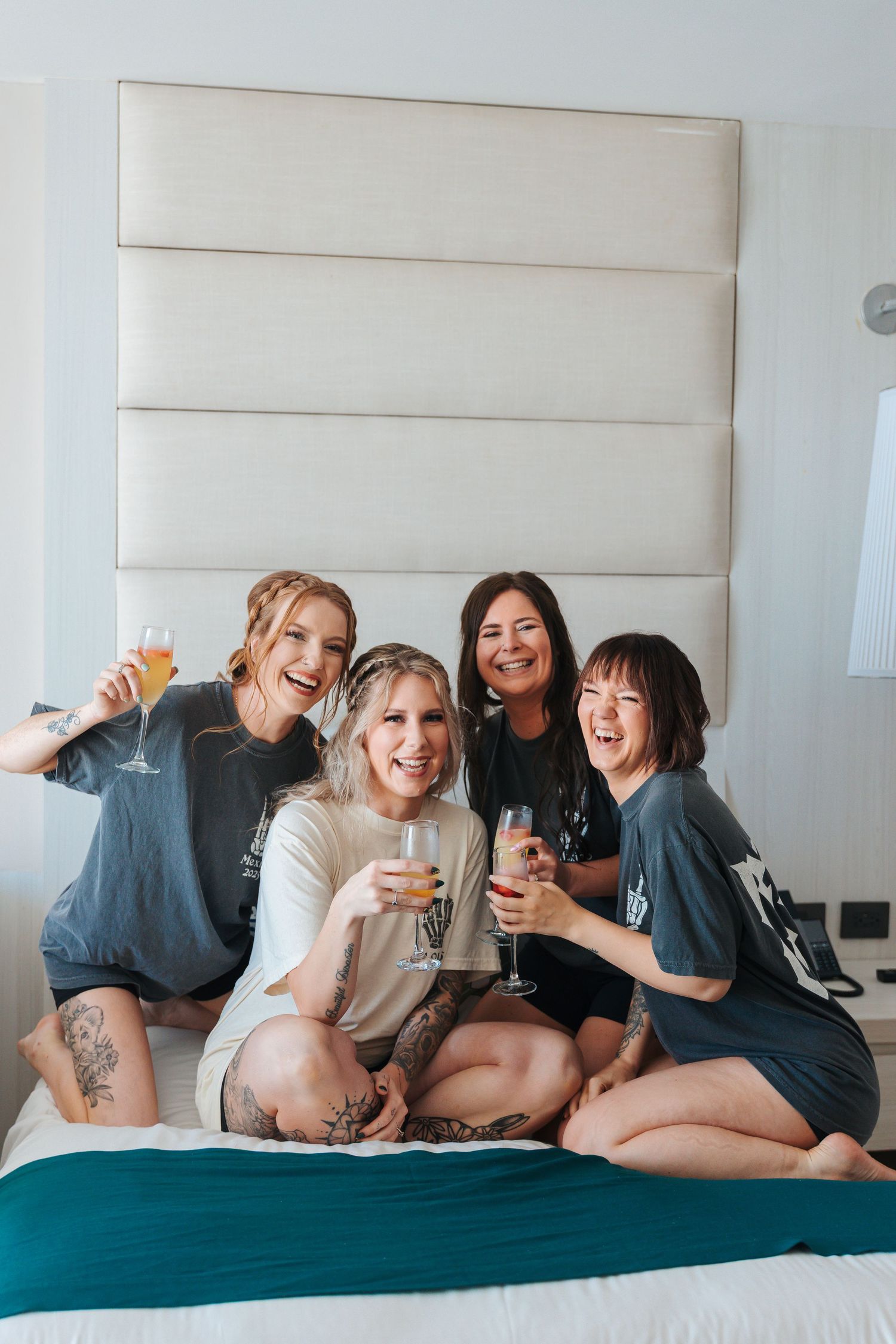 Four bridesmaidslaughing and holding champagne glasses while sitting on a hotel bed in matching gray shirts. mexico wedding resorts